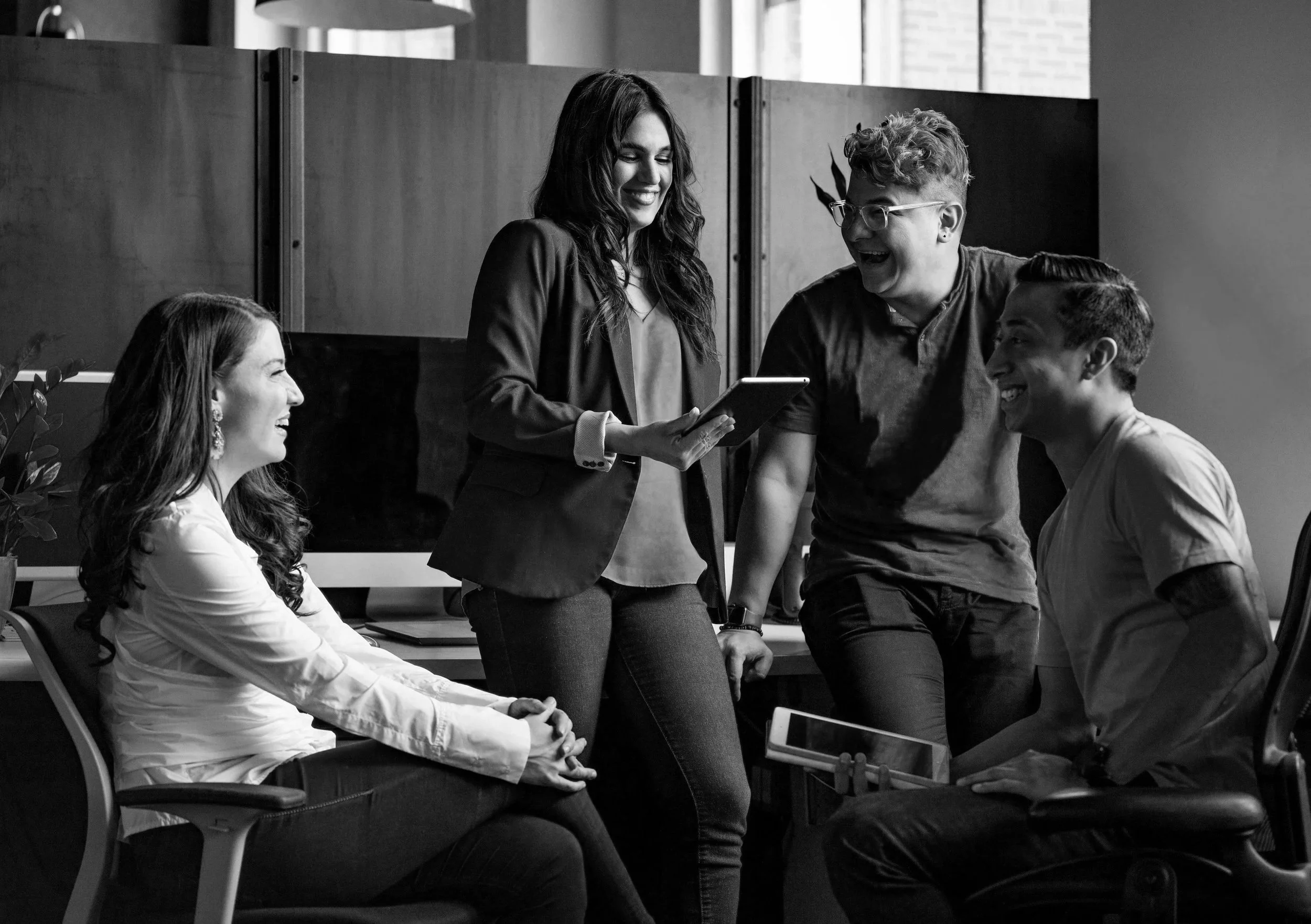 Four diverse coworkers in an office laughing and talking, one woman holding a tablet, others gathered around, relaxed and smiling.