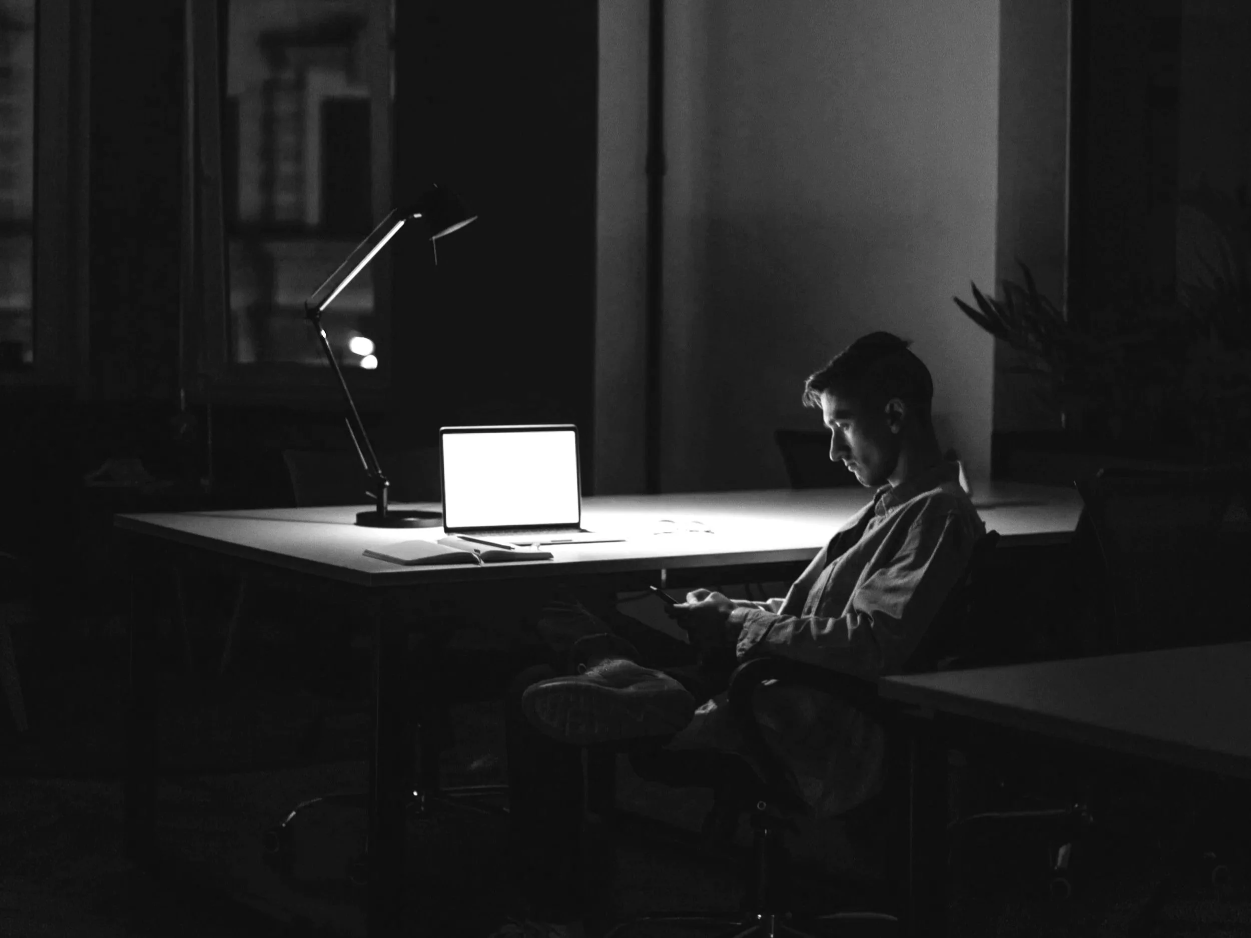 A man sitting at a desk in a dark room, illuminated by the light from a laptop screen, looking at his phone.