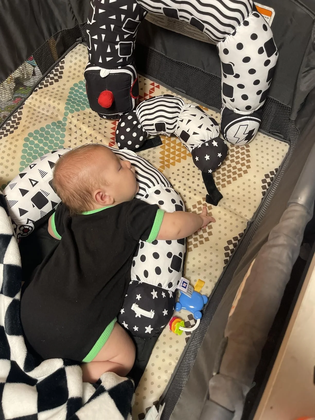 A sleeping baby lying in a colorful bed with a black and white geometric patterned blanket and plush toys, surrounded by a black and black-and-white toy arch with various patterns.