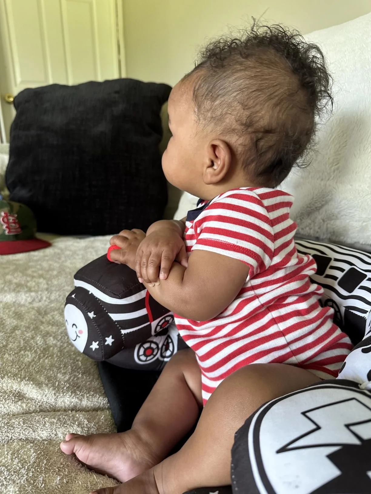A baby wearing a red and white striped shirt sitting on a bed, leaning over and holding a stuffed block with character illustrations, with a beige bedding and black pillows nearby.