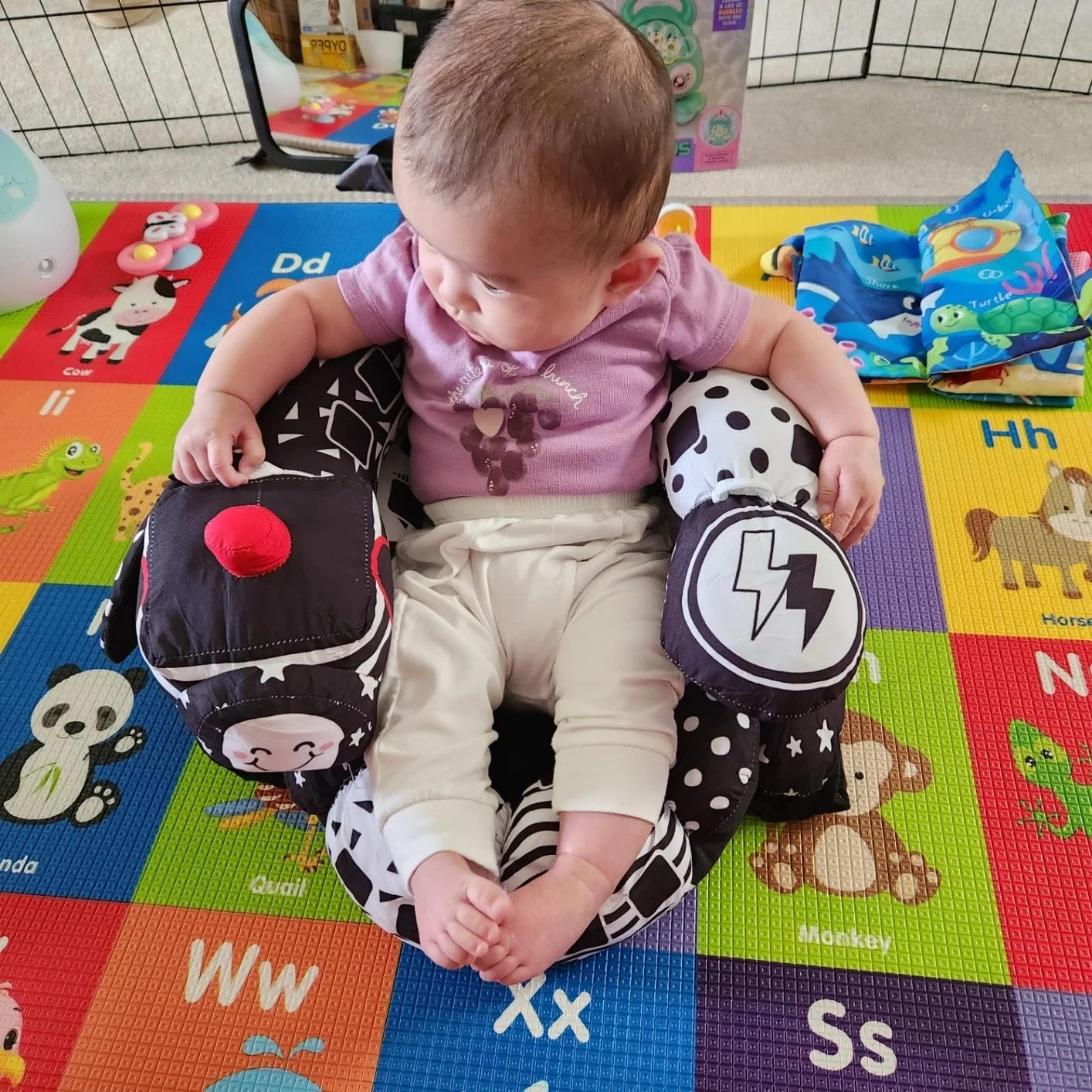 A baby sitting in a plush, decorated armchair placed on colorful alphabet foam mats with animal illustrations.