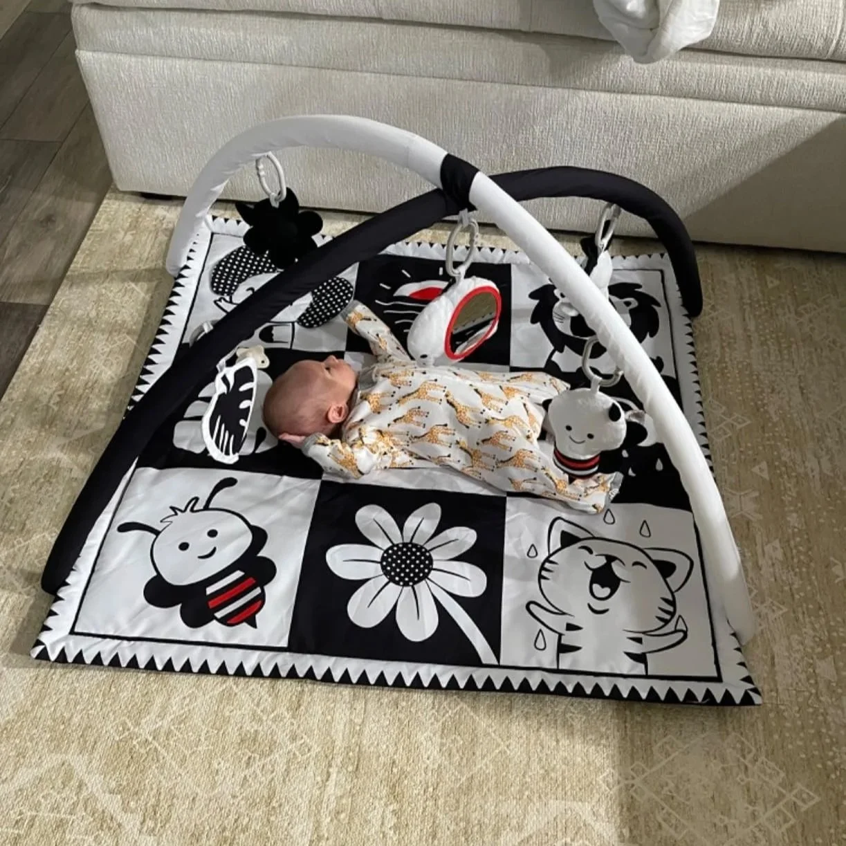 A baby lying on a black and white animal-themed playmat with hanging toys, in front of a white couch.