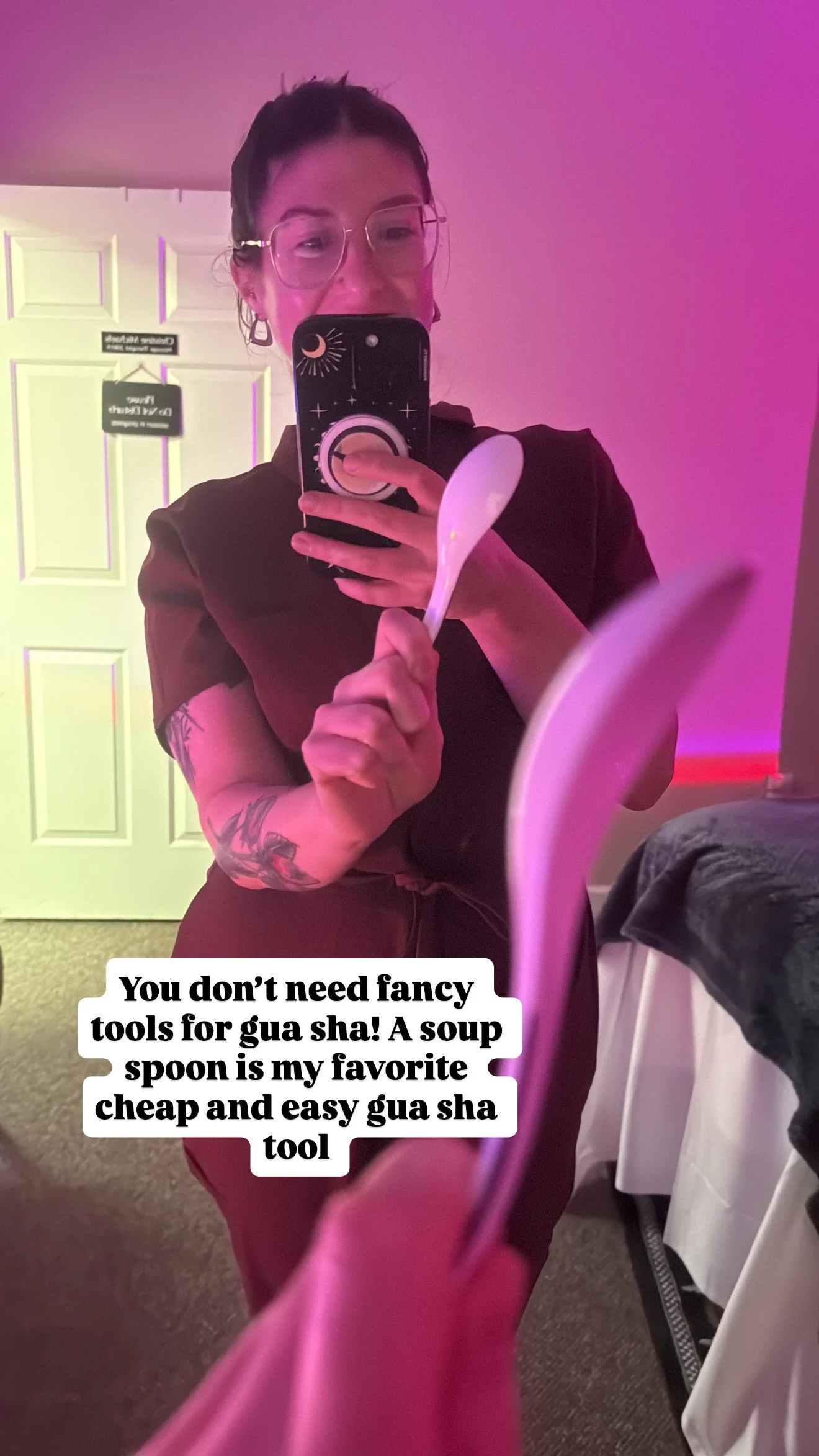 A woman with tattoos on her arm taking a selfie in a mirror, holding a soup spoon, with purple and pink lighting in the background. There are signs on a door behind her. A caption over the photo says, 'You don’t need fancy tools for gua sha! A soup spoon is my favorite cheap and easy gua sha tool.'