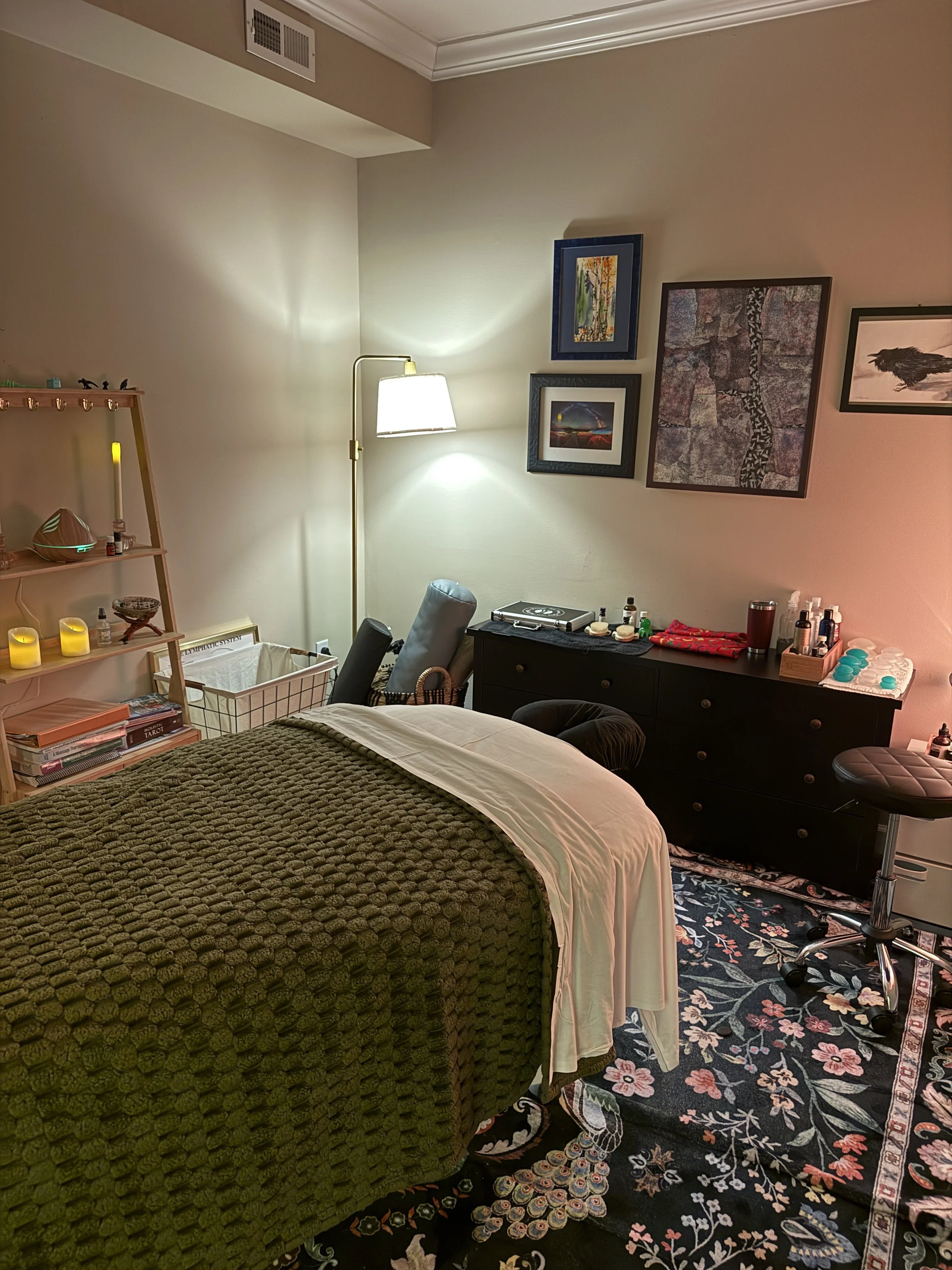 A cozy massage or therapy room with a bed covered in a textured green blanket, a black dresser with skincare or massage products, and framed artwork on the wall. There is a tall floor lamp providing warm lighting, and a black swivel chair near the dresser. A wooden shelf with candles and decor items is on the left, and a patterned rug covers the floor.