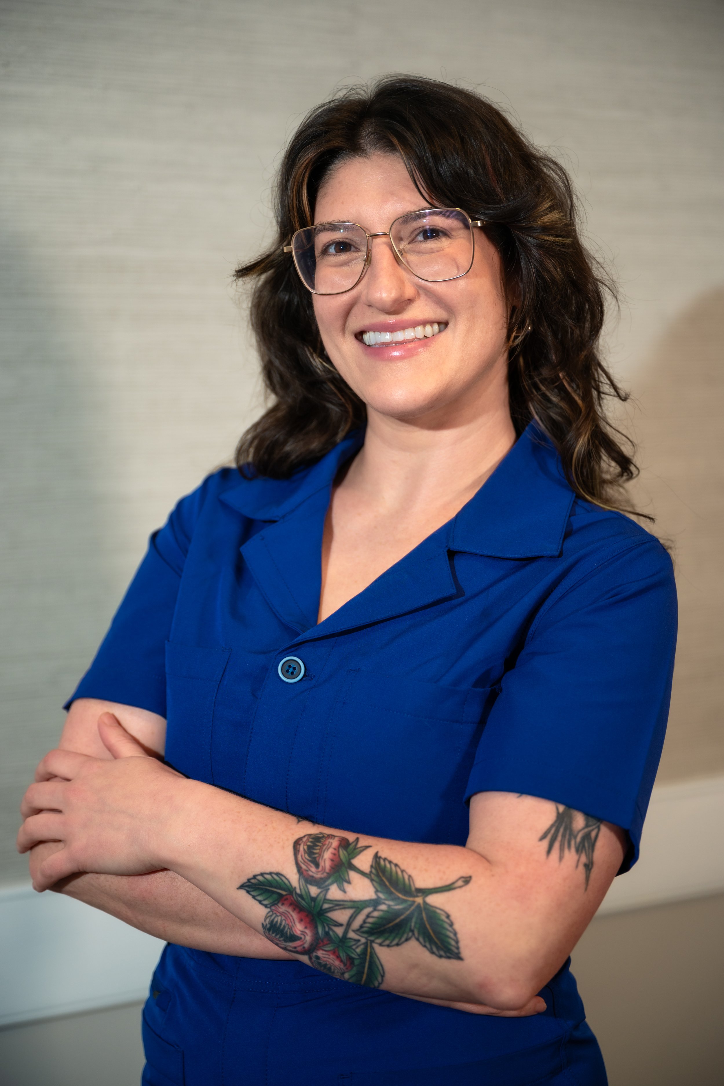 A woman with glasses and dark, wavy hair, smiling, wearing a blue shirt with her arms crossed, displaying a tattoo of strawberries and leaves on her right arm.