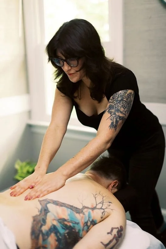 A woman with dark hair, glasses, and a tattoo on her arm is giving a massage to a man lying face down on a massage table, with large tattoos on his back. They are in a room with a window and soft natural light.