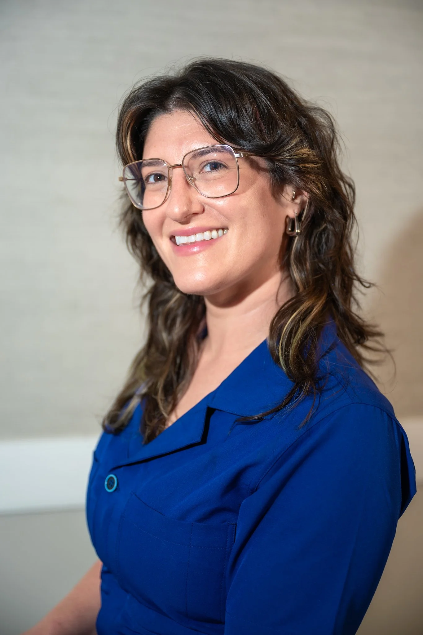 A woman with shoulder-length wavy brown hair, glasses, and earrings, smiling, wearing a blue shirt, standing in front of a light-colored wall.
