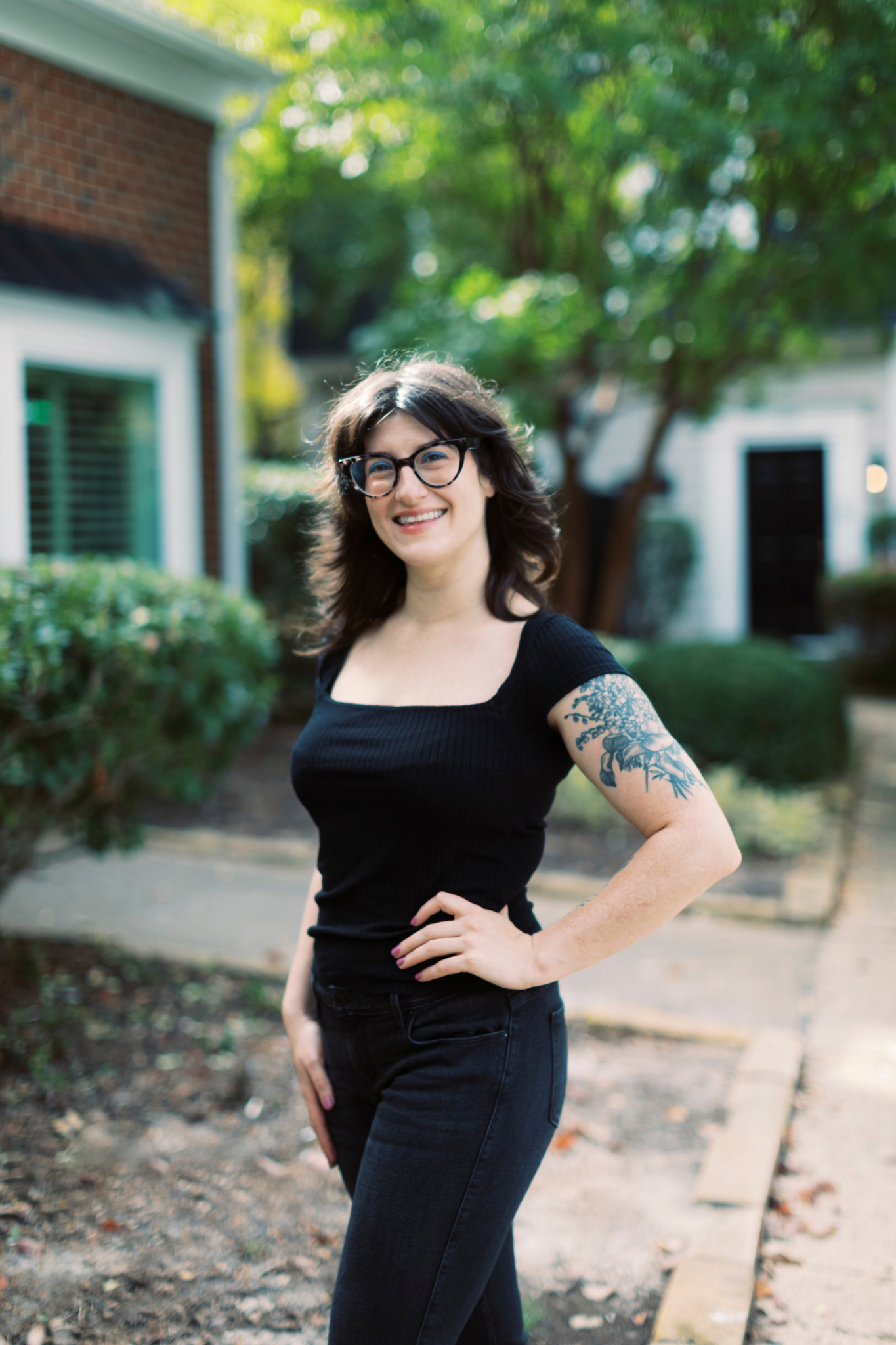 A woman with glasses, dark hair, and a tattoo on her arm, smiling outdoors in a residential neighborhood.