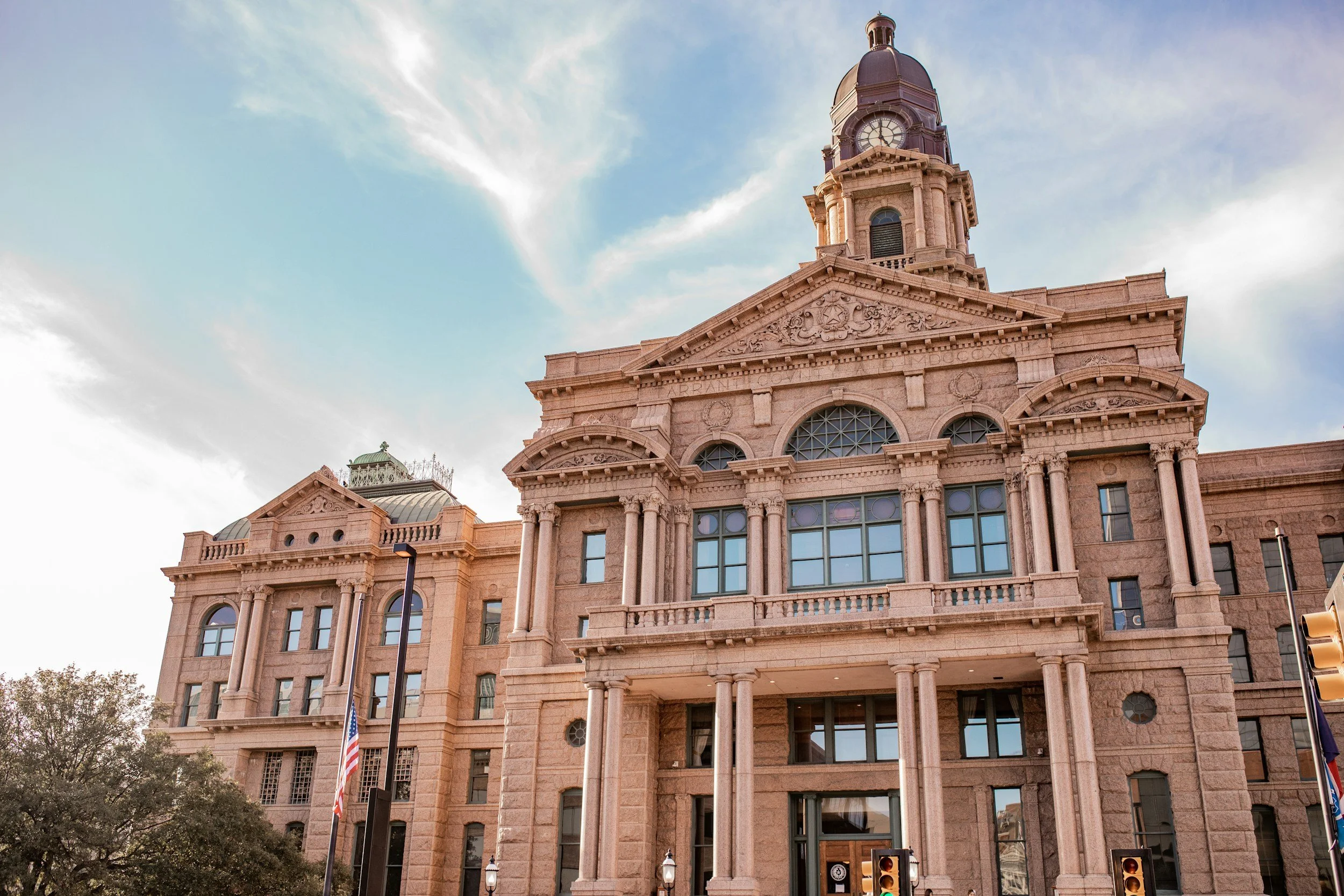 A historic pink stone courthouse with a clock tower against a partly cloudy sky. The building has large arched windows, decorative columns, and intricate stone carvings.