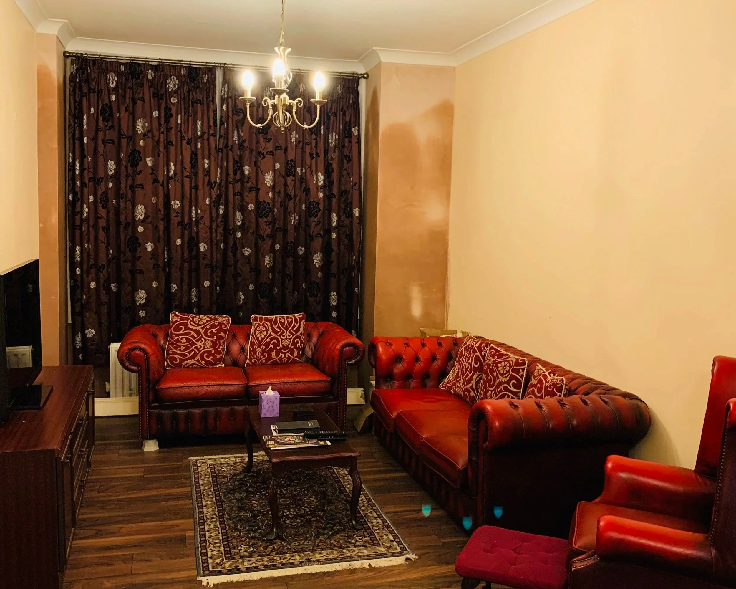 A before image of a living room that has red chesterfield sofas against a beige coloured wall, a mahogany coffee table is placed in the middle, brown patterned curtains adorn the window