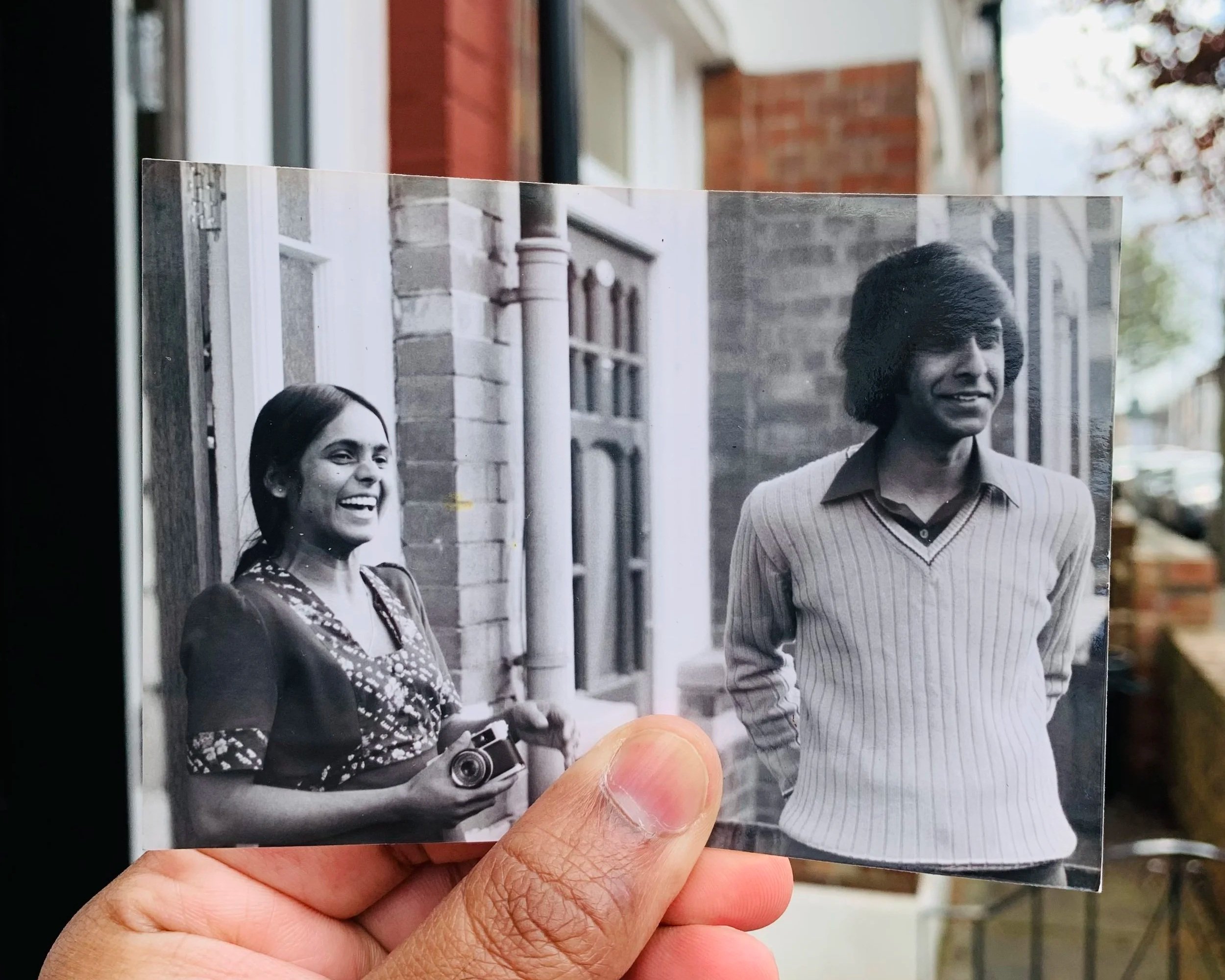 An old black and white image of a couple is being held over outside the front of a house, lining up to show the old and new together