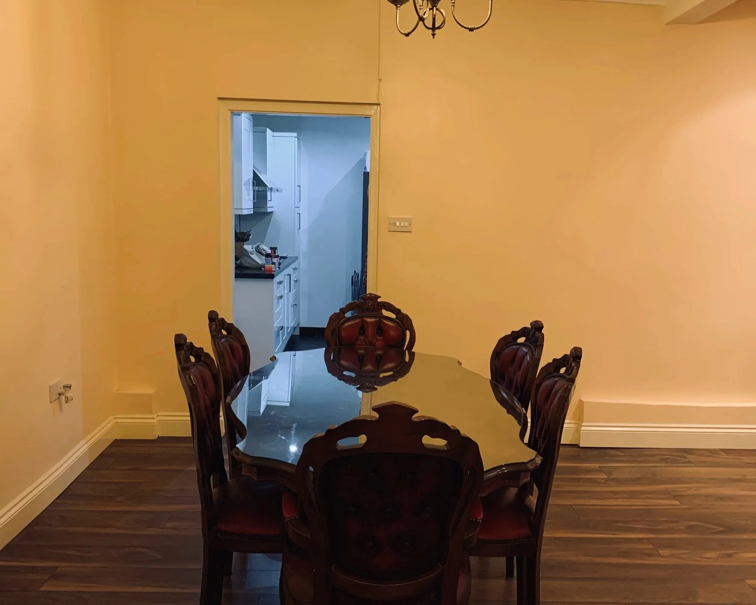 A before image of a dining room showing an old style dining table in the centre of the room, behind the entrance to a kitchen, the room is beige with brown wooden flooring