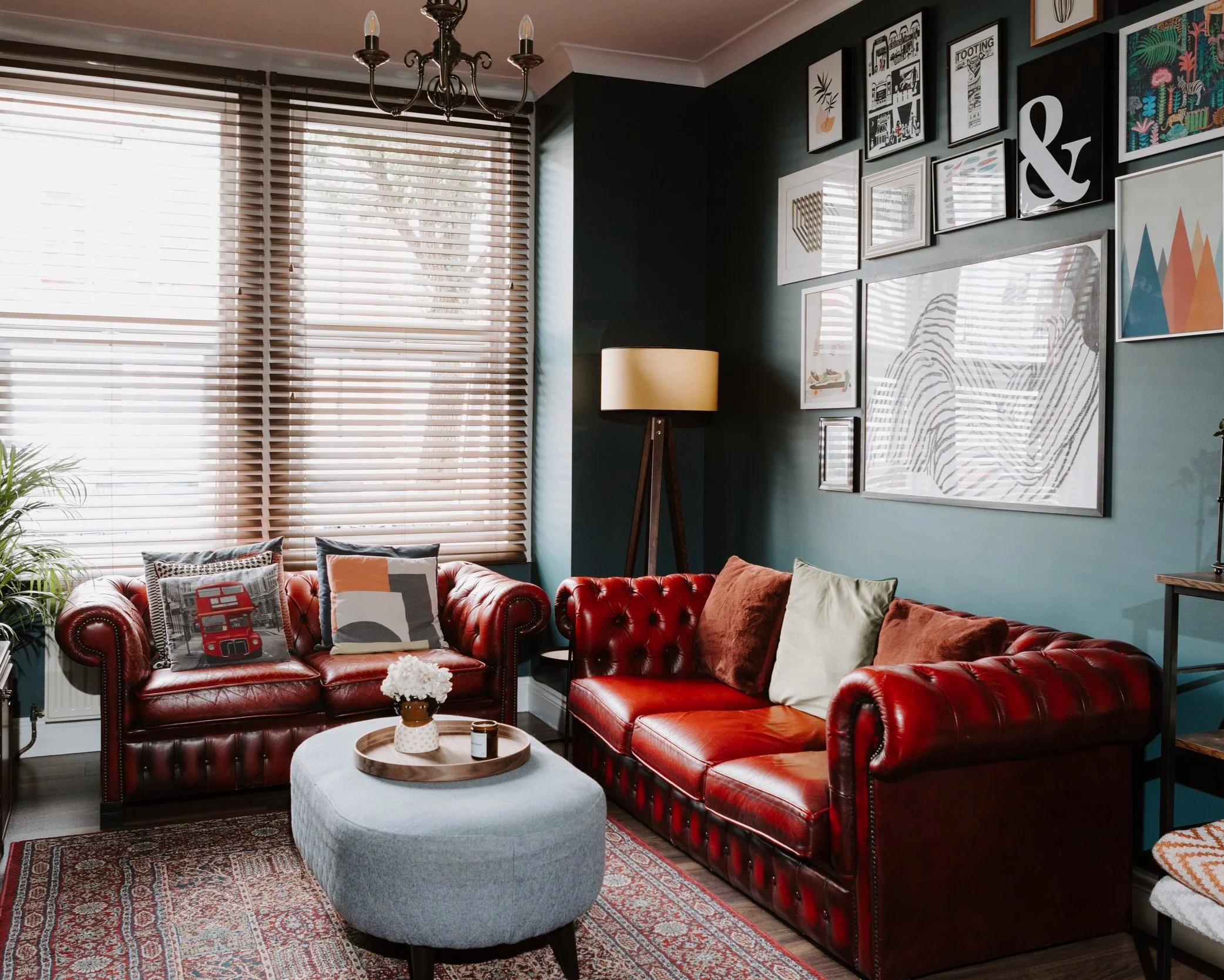 An after image of the same living room and red chesterfield sofas are now in an updated designed space, teal green walls with a gallery of framed pictures and prints, the space is decorated further with a yellow lamp and grey futon style piece that i