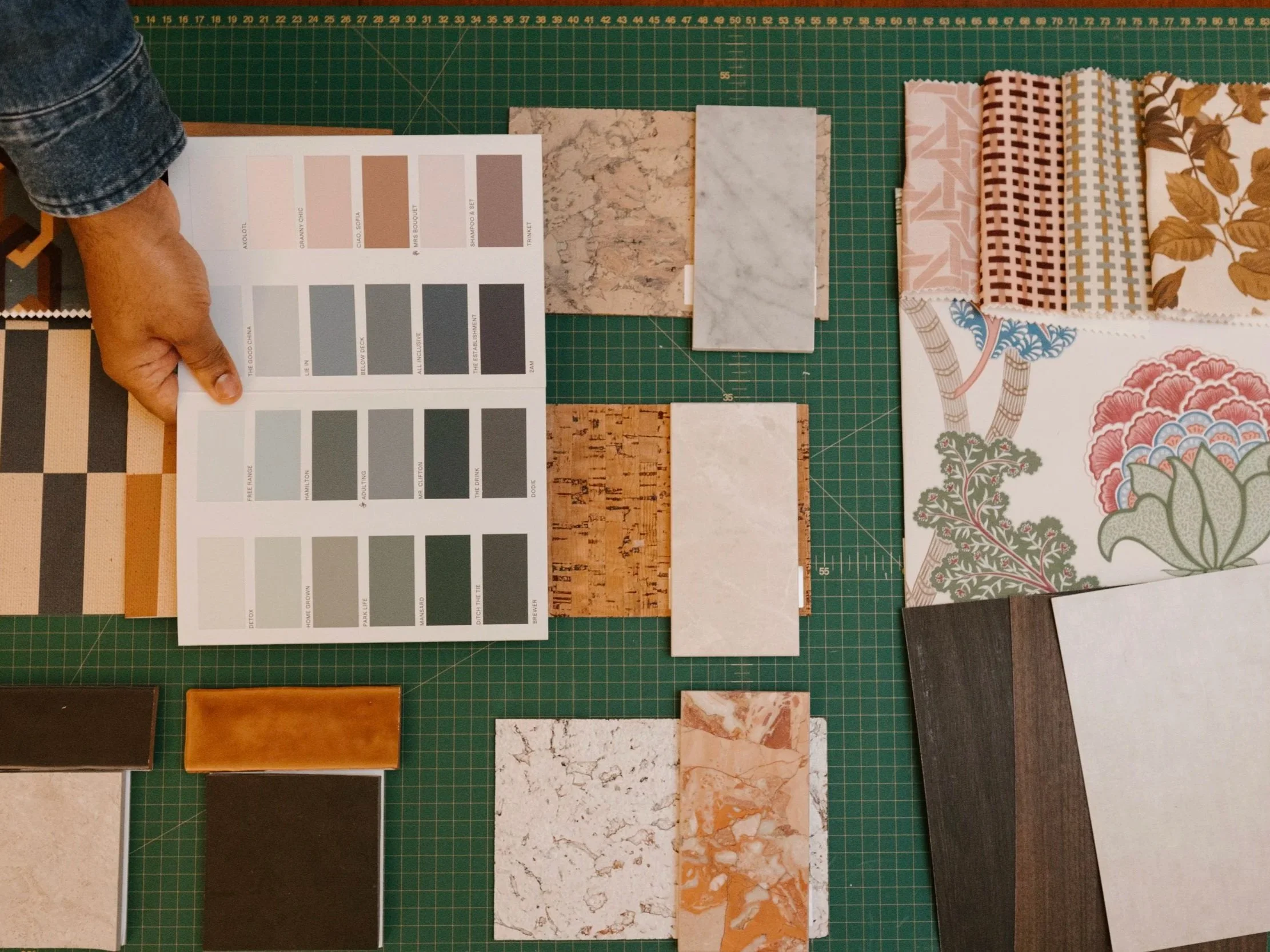 A person holding a color sample chart at a workspace with various tiles and fabric samples arranged on a green cutting mat.