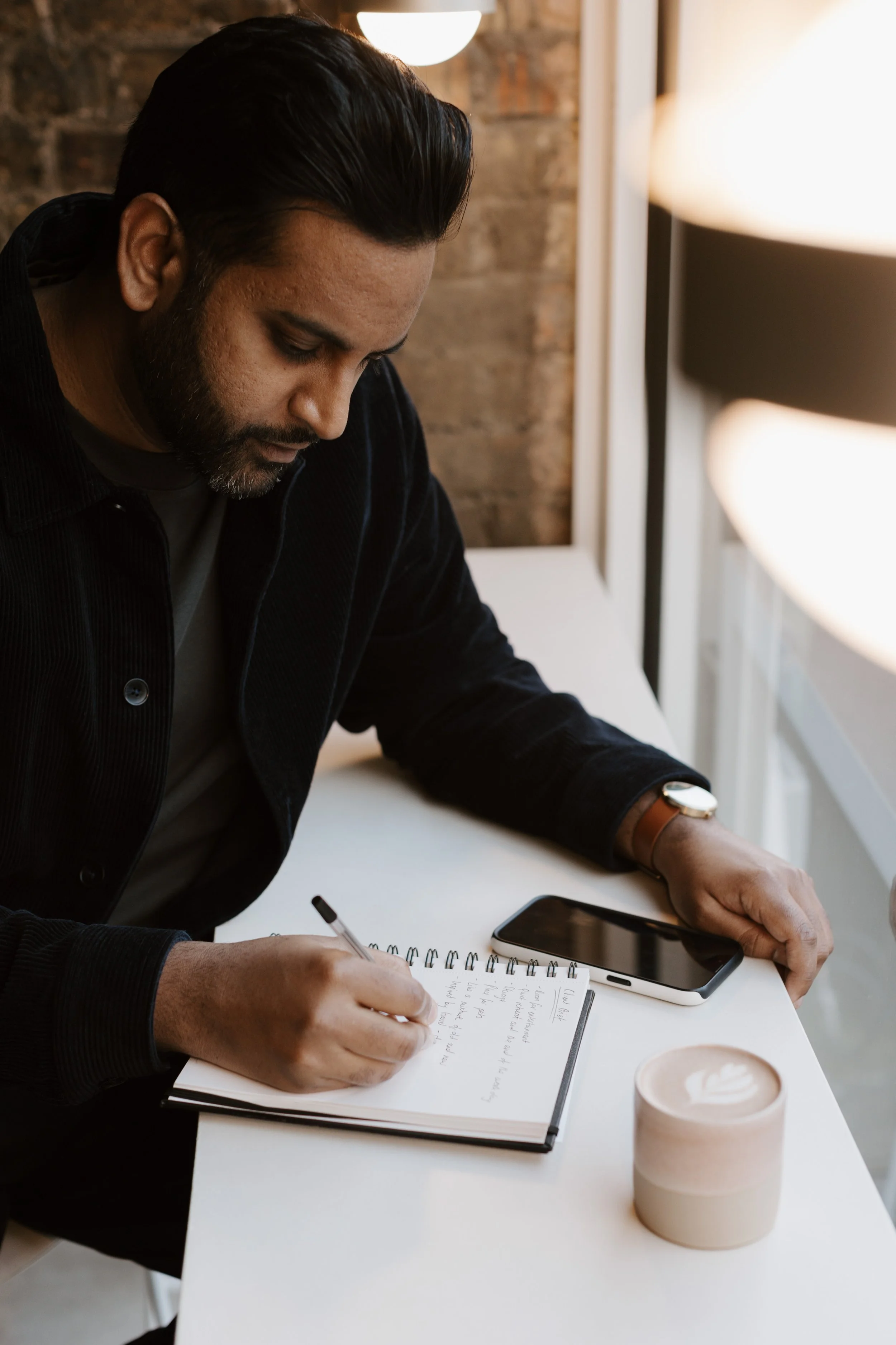 A man with dark hair and a beard, sitting at a white desk, writing notes in a notebook with a black pen. He is wearing a dark jacket and a watch on his left wrist, with a smartphone and a cup of coffee nearby. The background has a brick wall and a large window.