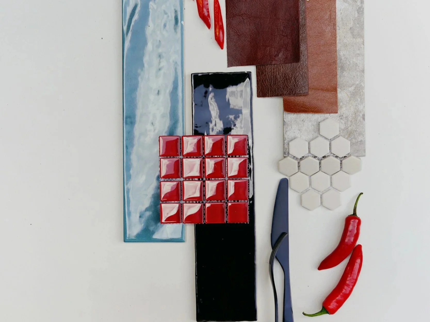 Various materials and objects arranged on a white surface, including red peppers, small red tiles, hexagonal white tiles, leather or faux leather swatches in brown and other shades, a rectangular blue and white patterned tile, a black piece, a small rectangular black and blue item, and a blue-handled cutting tool.