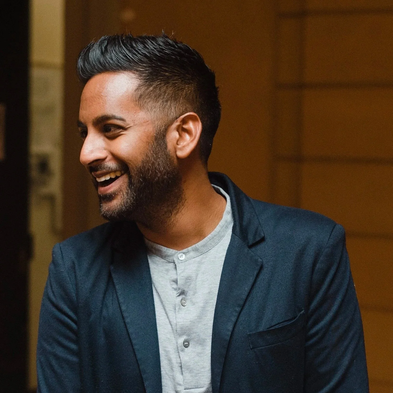 A man with short dark hair and a beard, smiling and looking to his left, wearing a light gray shirt and a dark blazer, indoors with warm lighting.