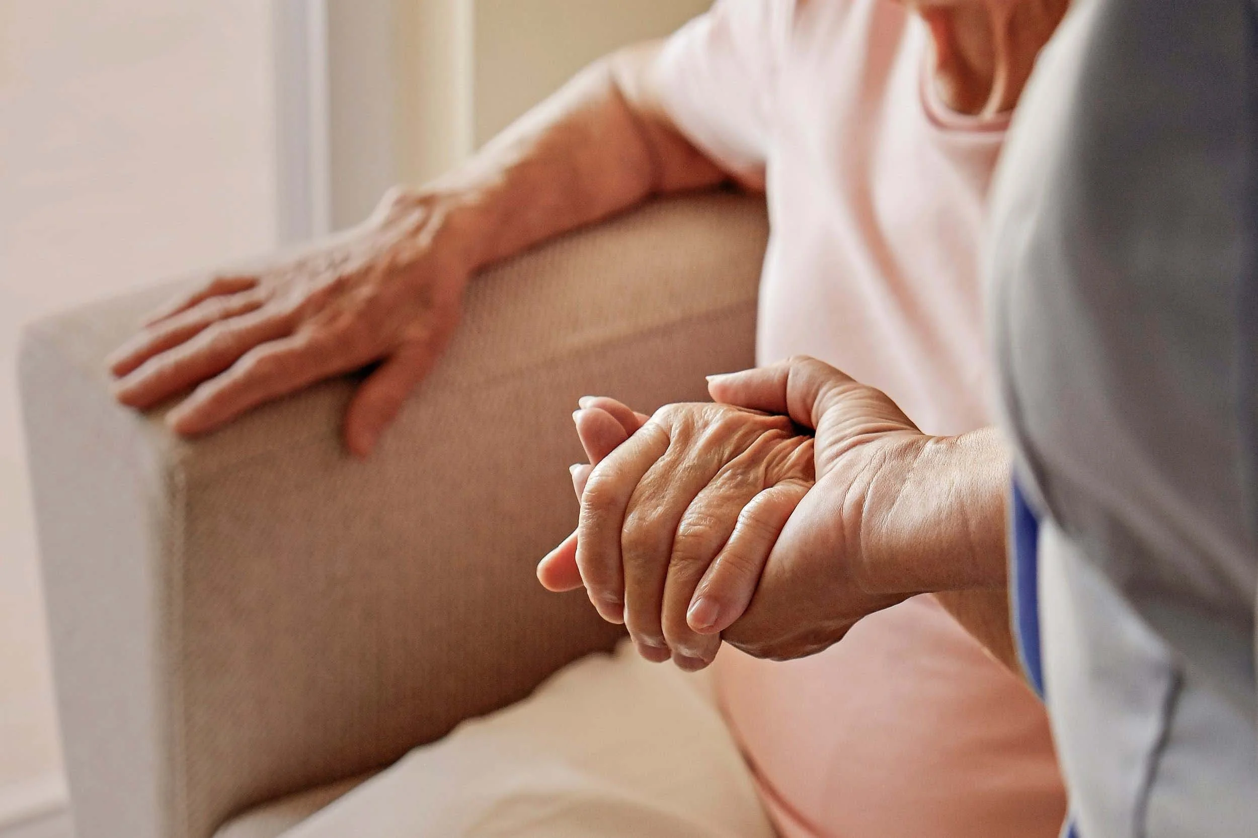 A personal care assistant holding an elderly woman's hand, with the elderly woman resting her arm on the back of a beige sofa.
