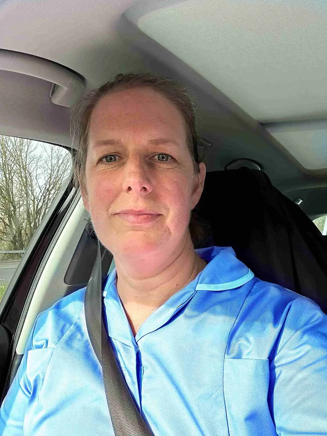 Woman with short brown hair and blue eyes taking a selfie inside a car, wearing blue scrubs and a seatbelt, with a window showing trees outside.