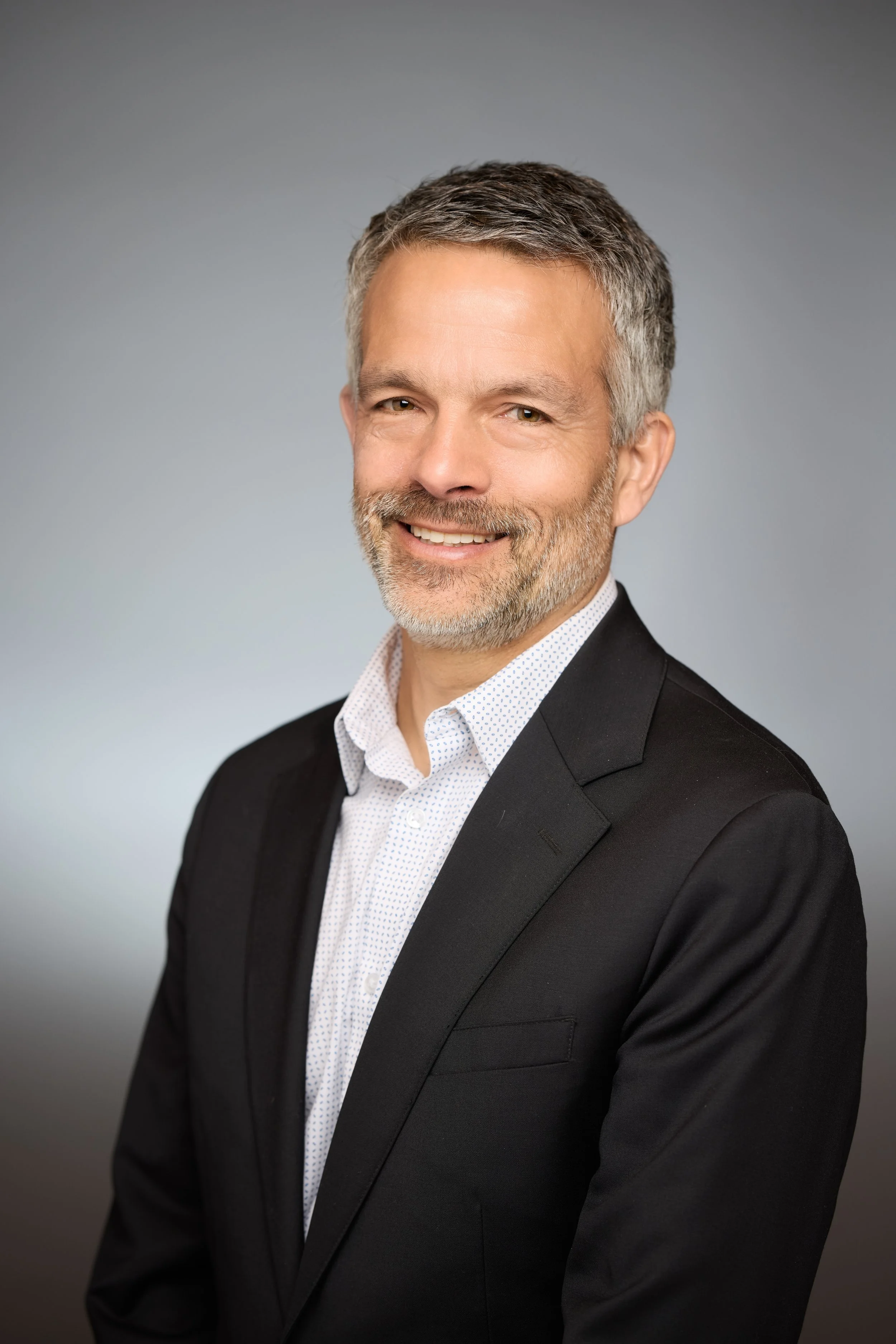 Professional headshot of a middle-aged man with gray hair and beard, wearing a black suit jacket and white dress shirt, smiling against a neutral gray background.
