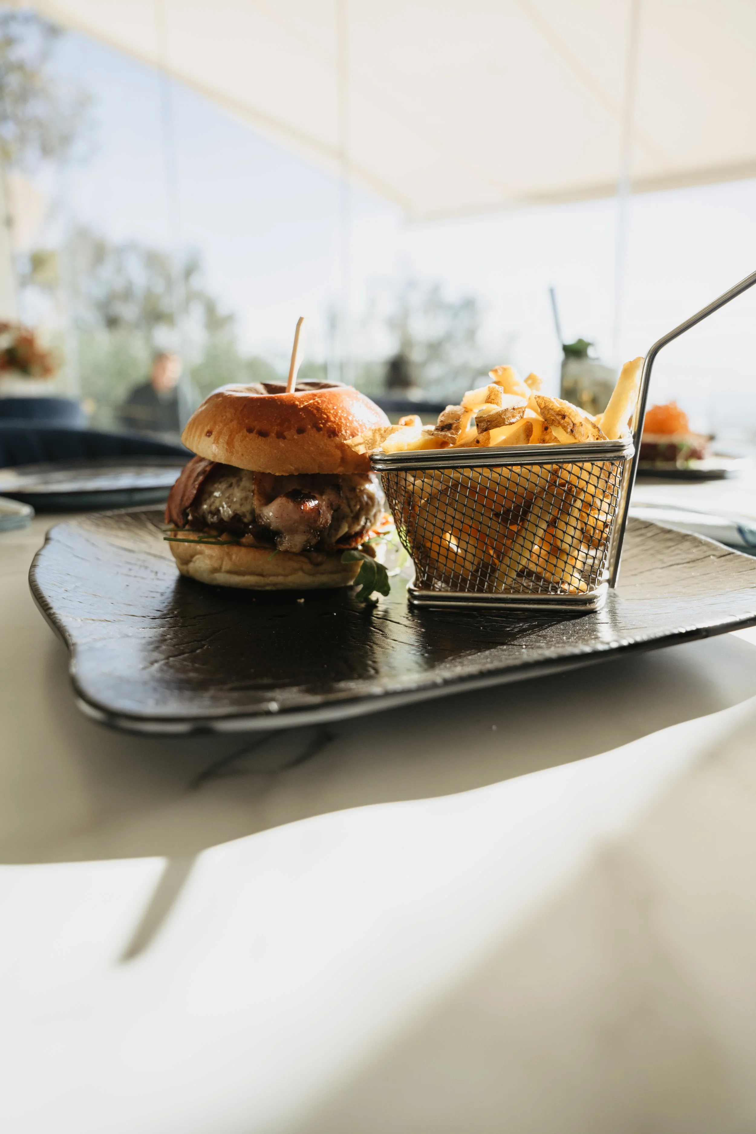 A cheeseburger with a bun, lettuce, patty, and cheese, served with a side of French fries in a small metal basket, on a dark serving tray at a restaurant.