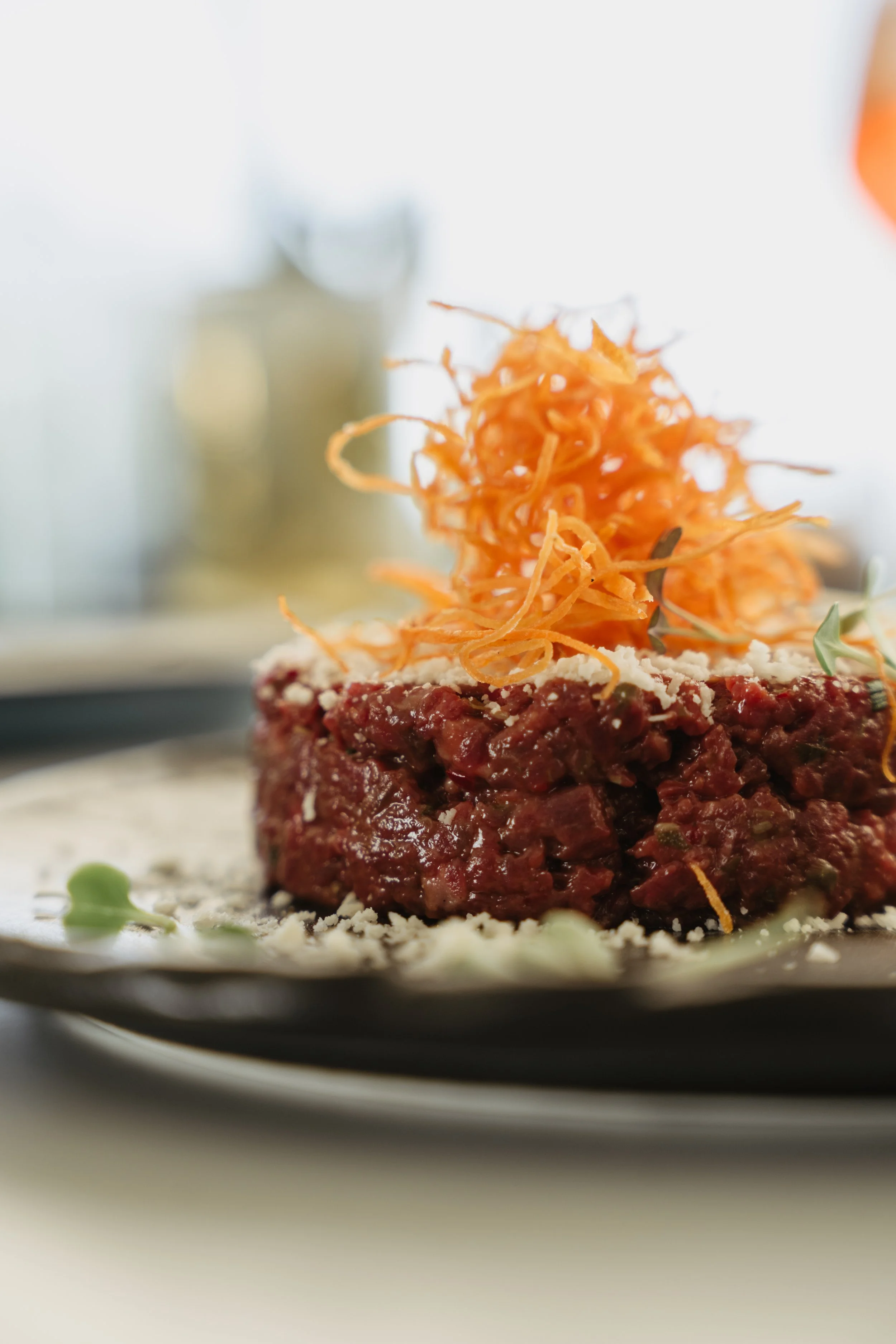 Close-up of a raw beef steak topped with grated cheese and crispy shredded carrots, garnished with microgreens, on a black plate.