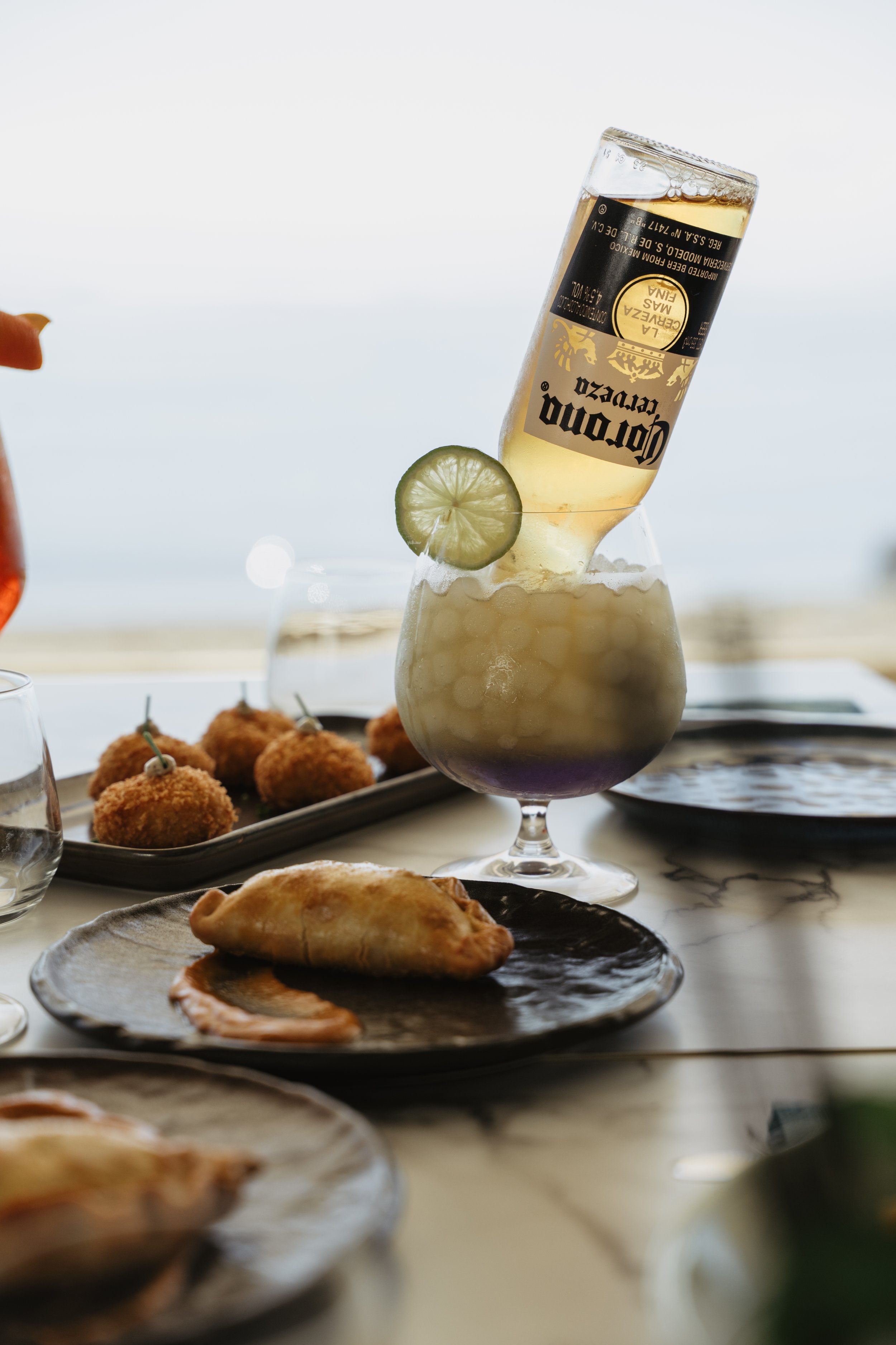 A drink with a lime slice and a Corona beer bottle in a glass, with fried appetizers on plates on a table near a beach.