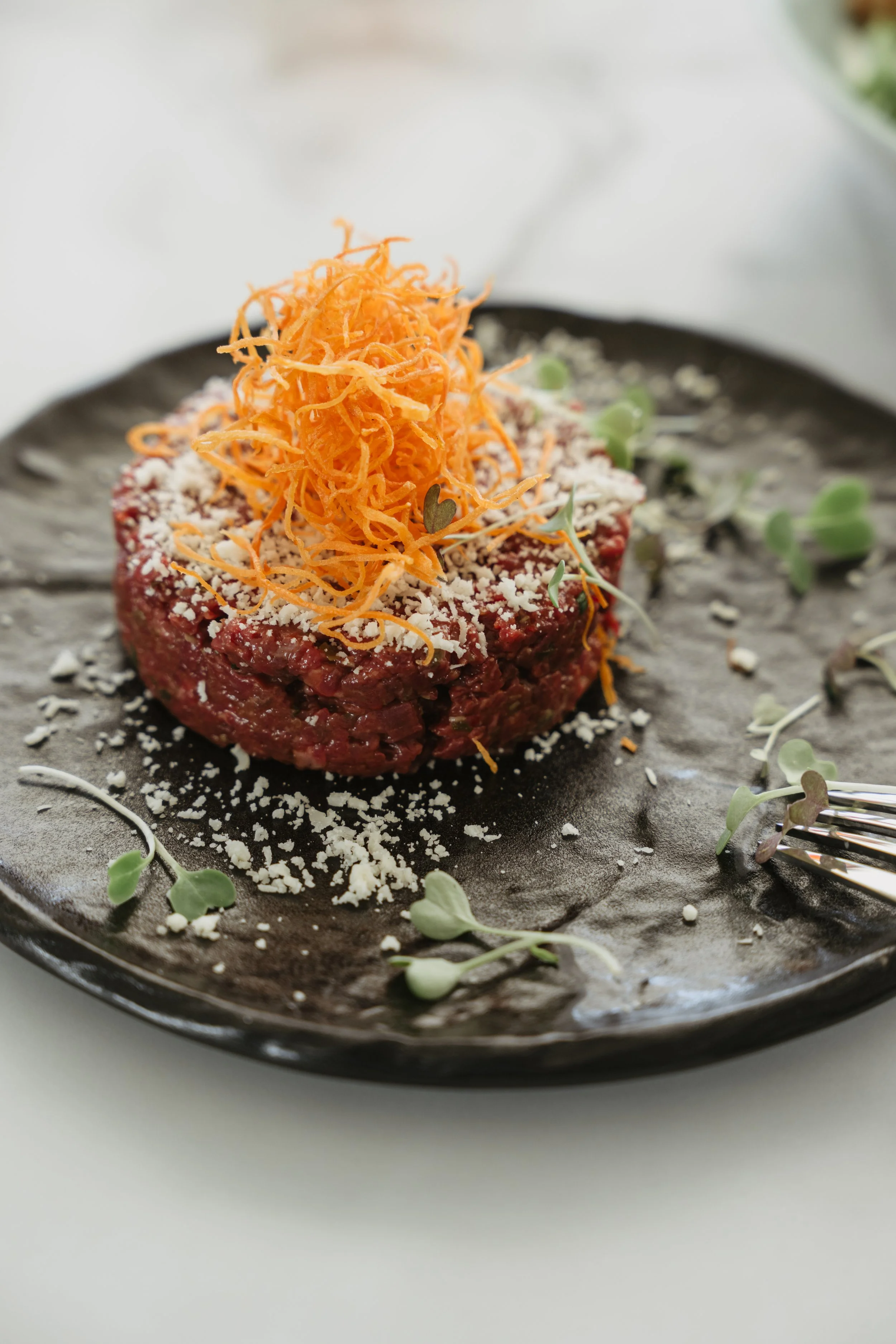 Raw beef tartare topped with shredded carrots and microgreens on a black plate.