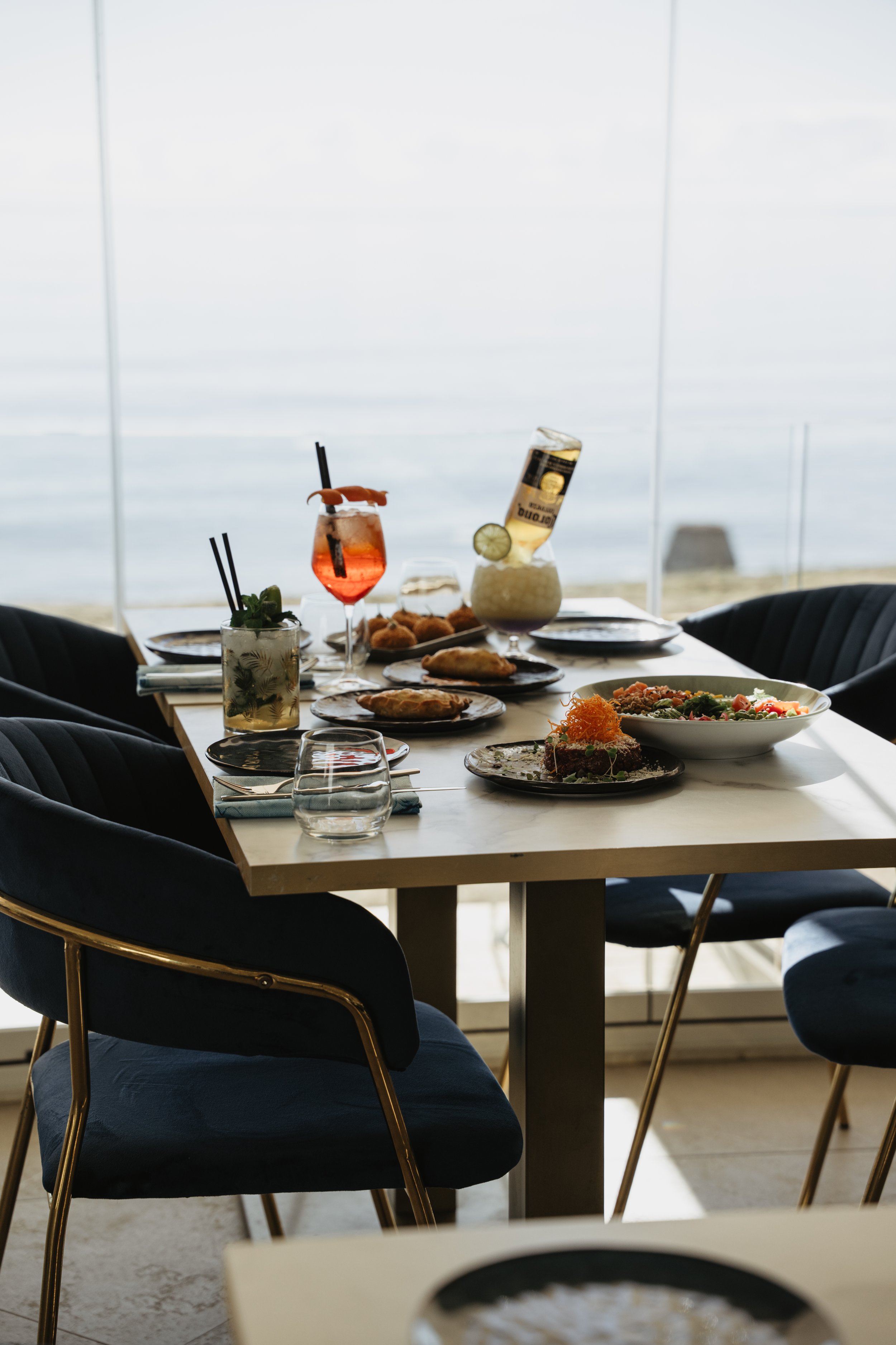 A table set with various dishes and drinks, overlooking a window with a view of the ocean.
