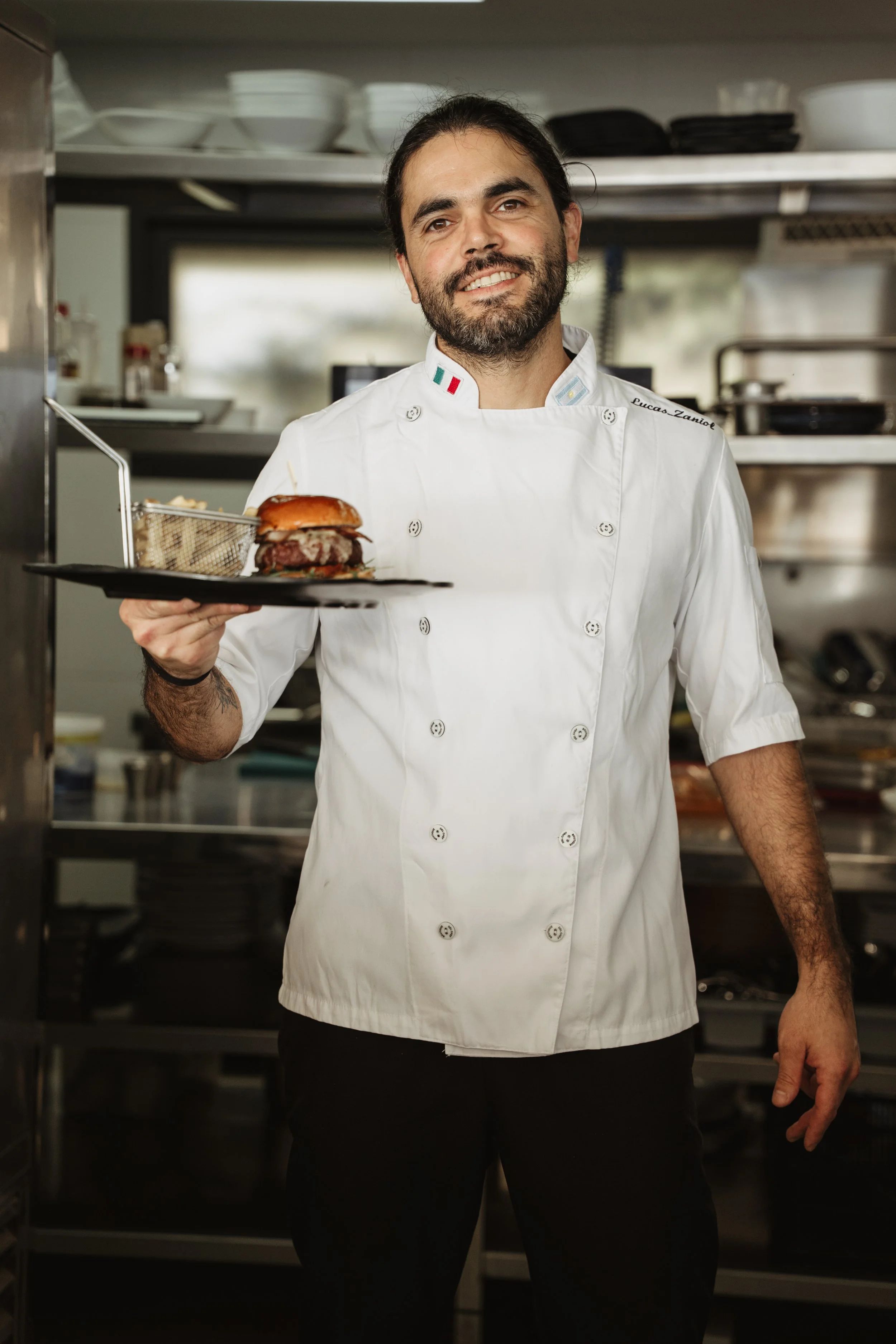 A male chef in a white uniform holding a plate with a burger and fries, smiling in a commercial kitchen.