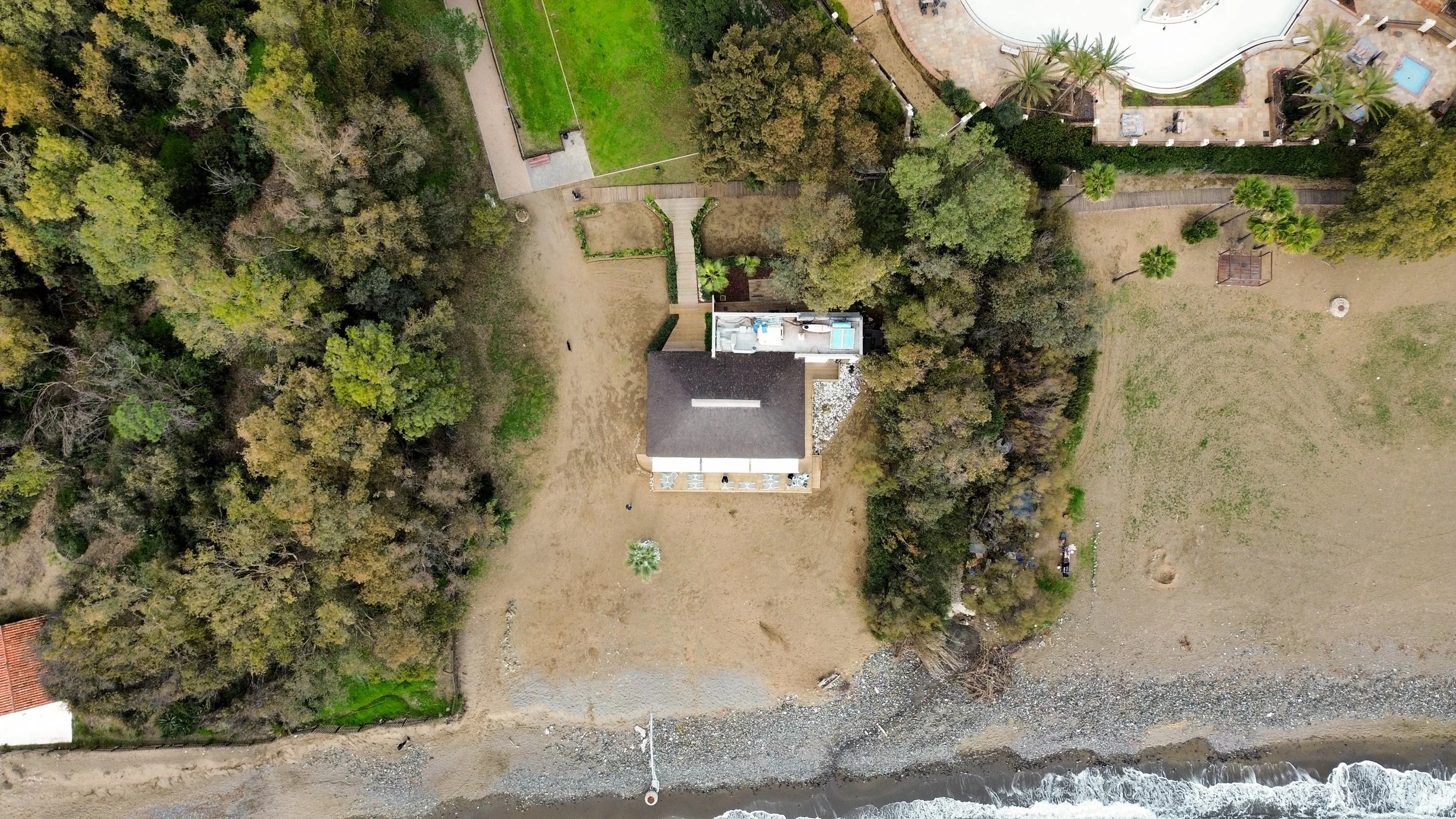 Aerial view of a house near the beach, with trees around and a lawn, adjacent to a rocky shoreline with ocean waves.