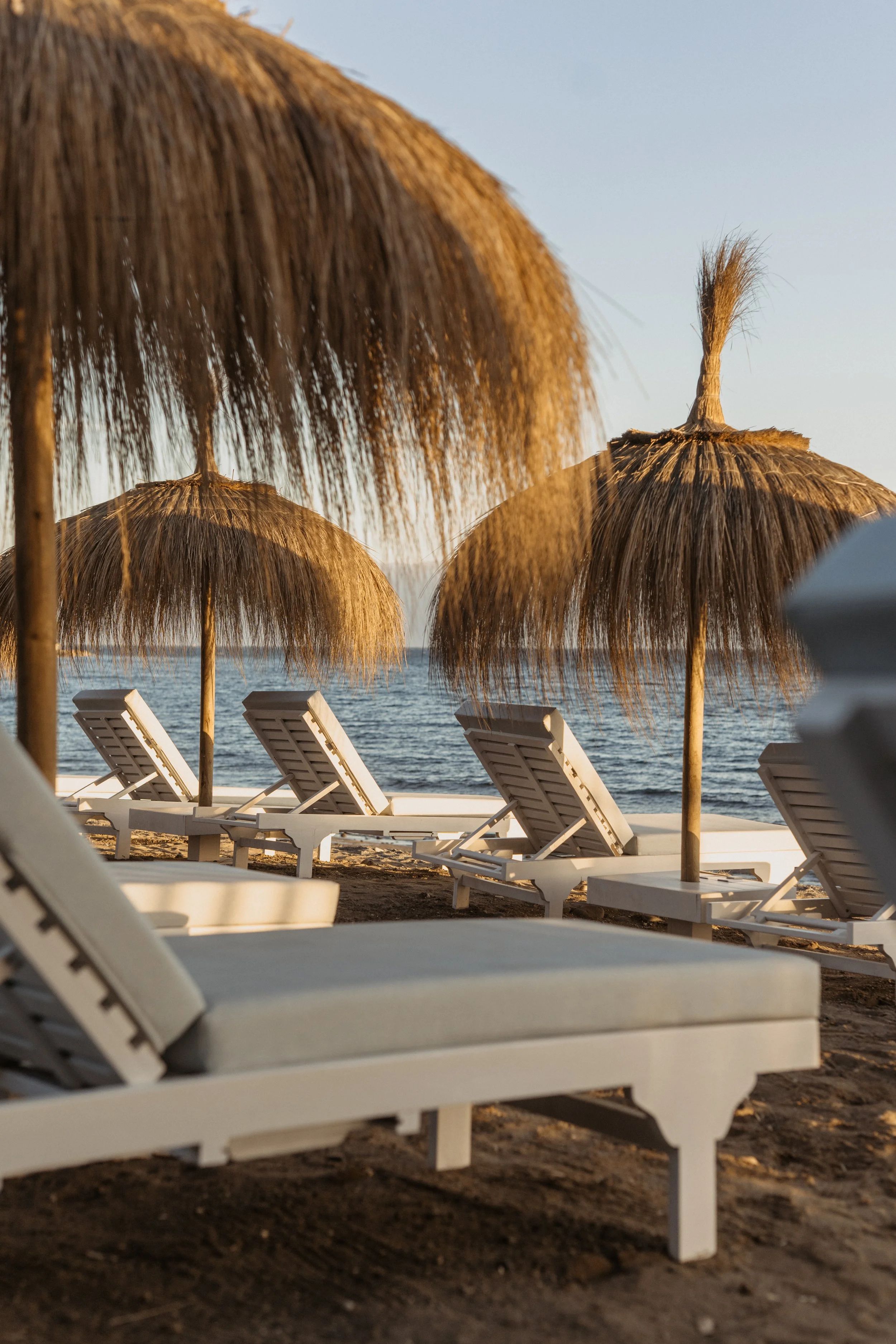 Beach scene with white lounge chairs under thatched umbrellas facing the ocean at sunset.