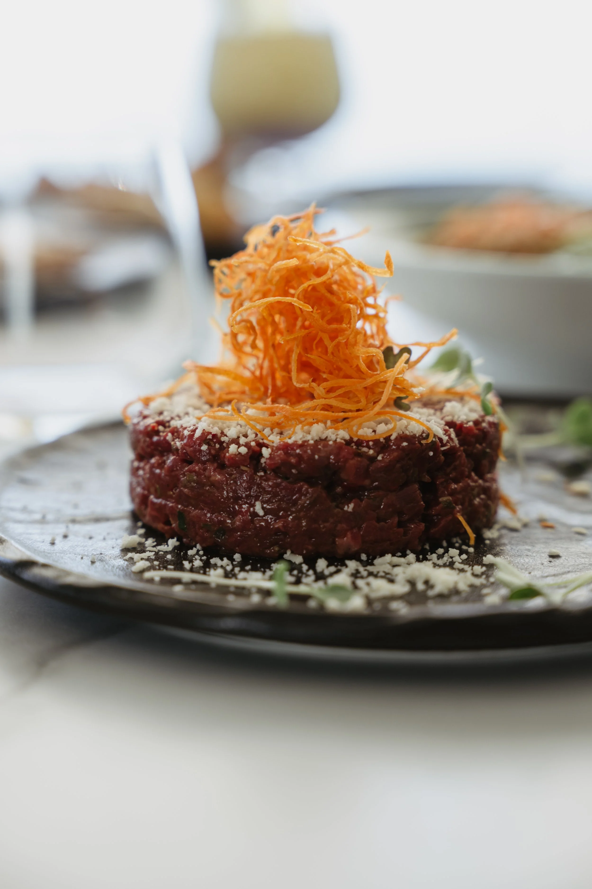 Close-up of a raw beef patty topped with shredded cheese and crispy fried onions, garnished with microgreens, served on a dark plate.