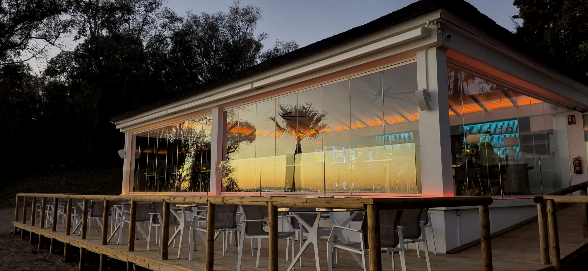 A modern restaurant with glass walls reflecting a sunset and a palm tree, surrounded by a wooden deck with white chairs and tables.