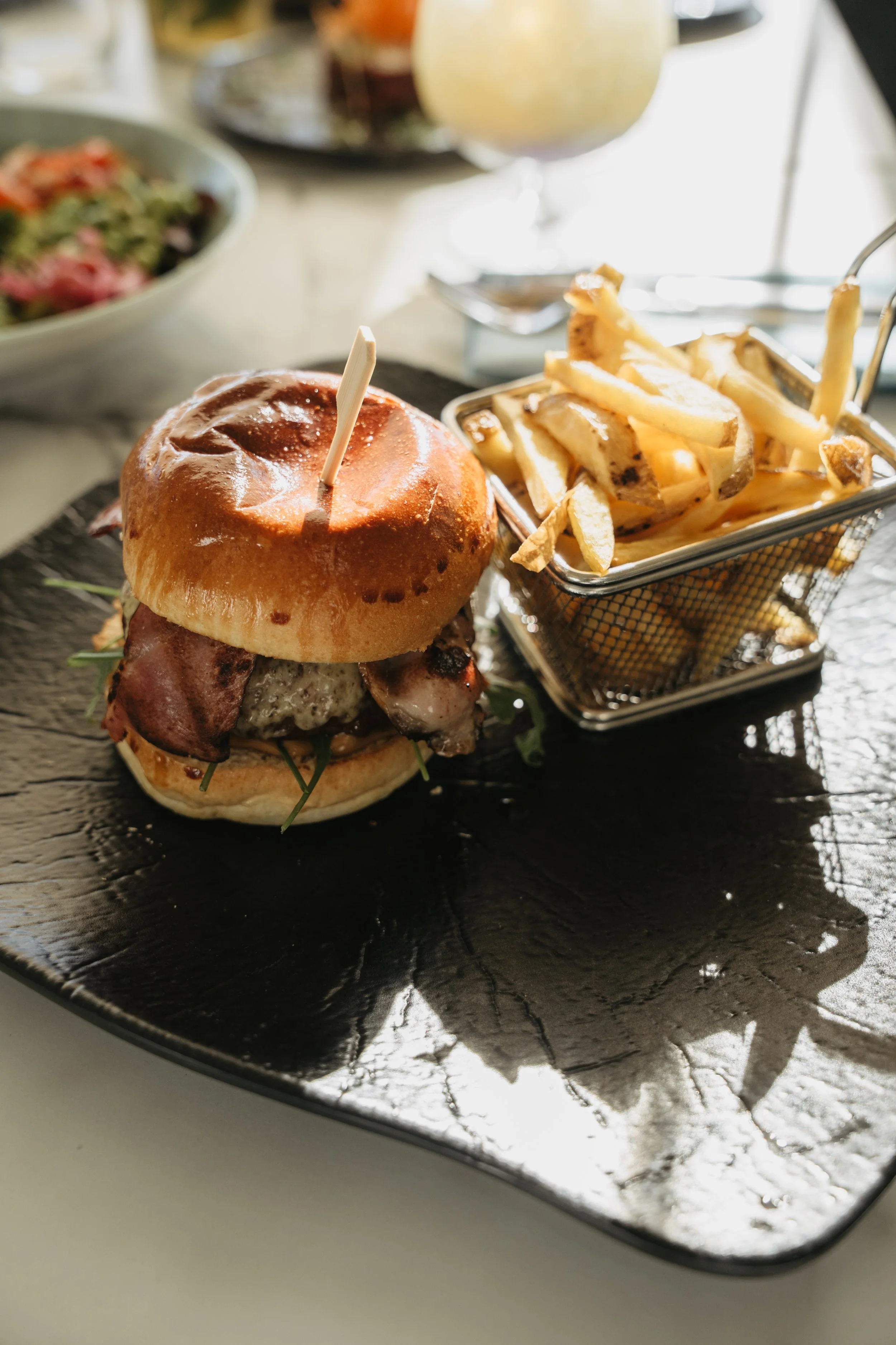 A burger with bacon and greens served with a side of French fries on a black plate.