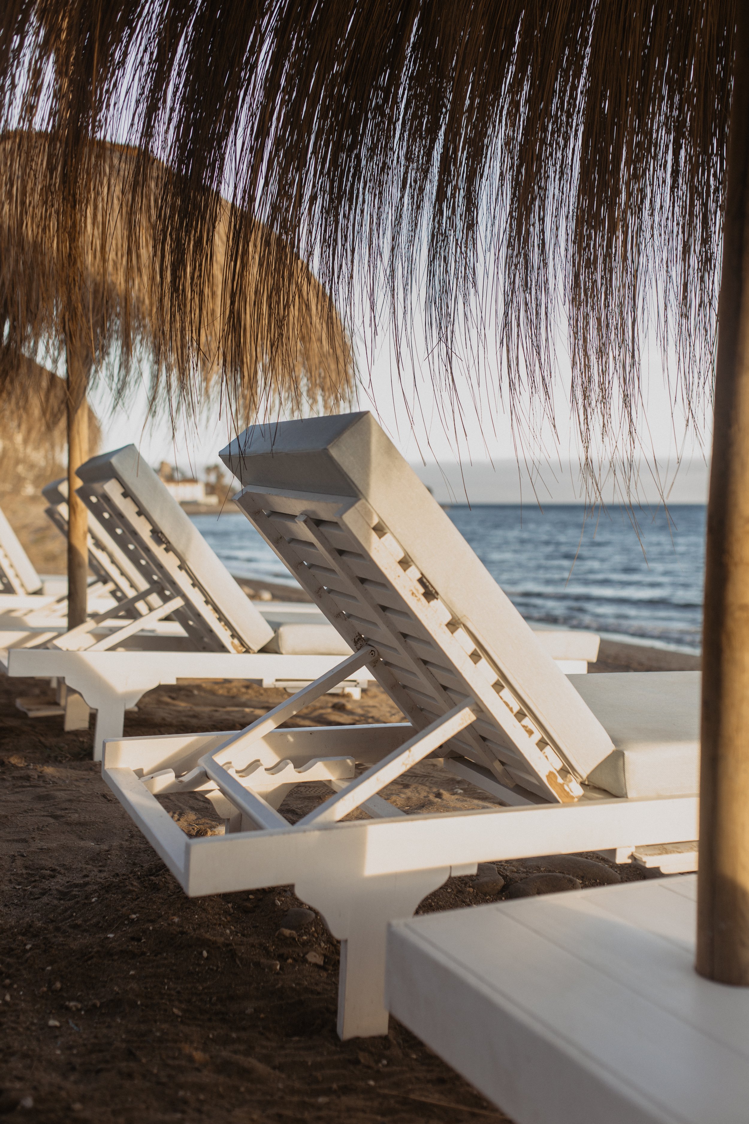 Empty white lounge chairs on a sandy beach with thatched umbrellas and the ocean in the background.