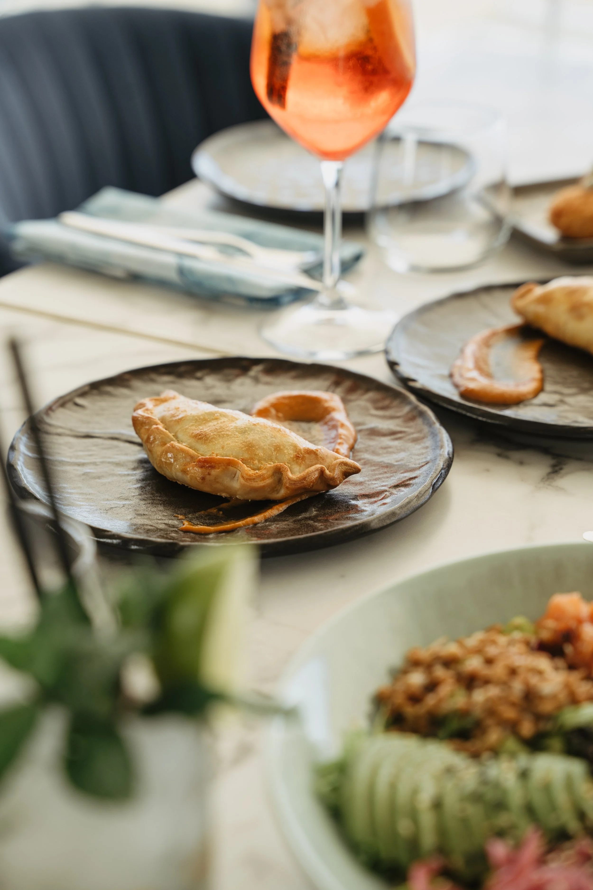 A plate with a cooked dumpling and a slice of roasted vegetable on a black ceramic plate, set on a table at a dressed dining setting with a wine glass and additional plates visible.