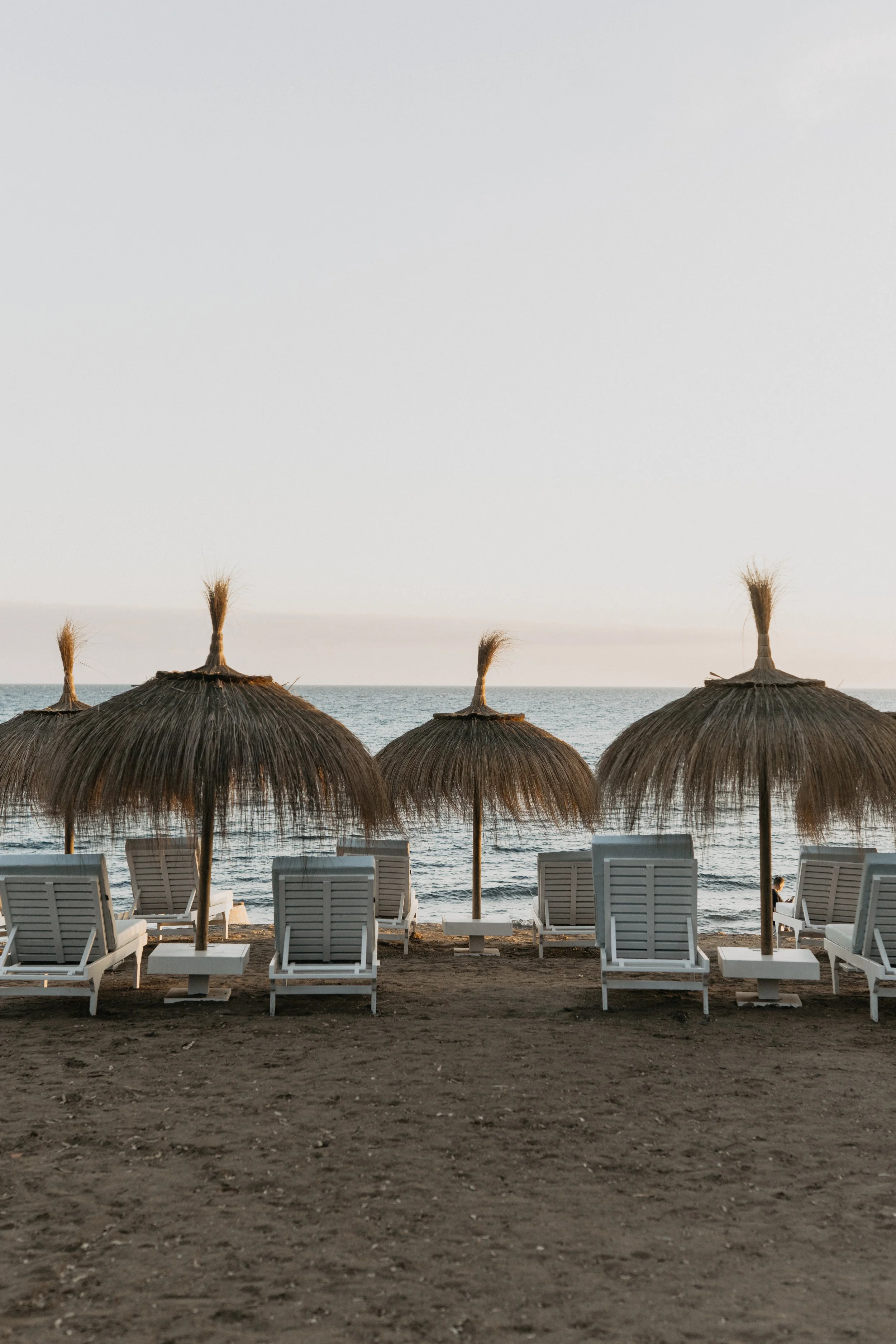 Rows of empty lounge chairs under straw beach umbrellas on a sandy beach facing the sea with a peaceful, clear sky.