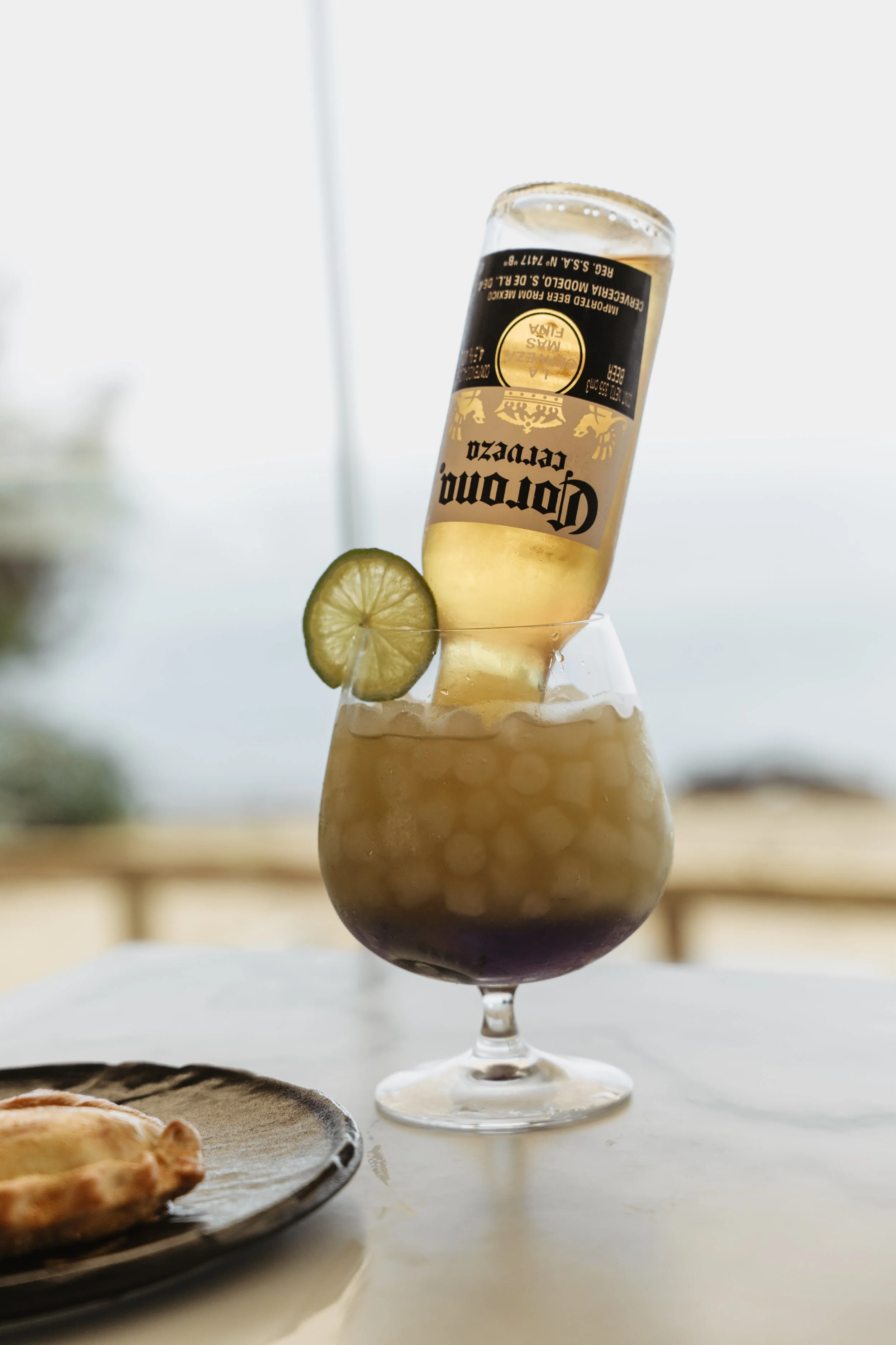 A glass of mixed drink with a lime garnish, topped with a beer bottle balanced on the rim, and a partially visible pizza on a plate in the foreground.