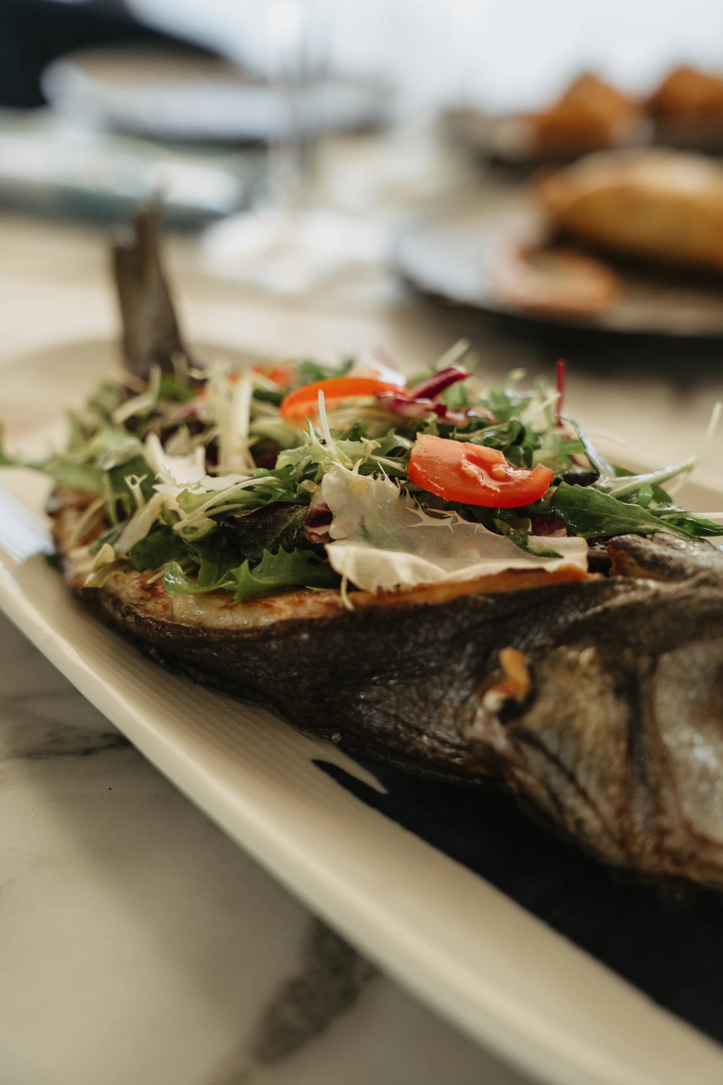 Baked fish topped with mixed greens, cherry tomatoes, and cheese on a white platter.