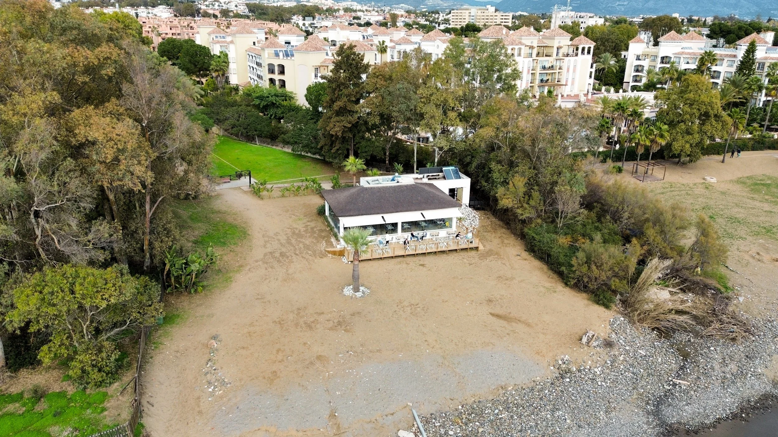 Aerial view of a beachside house with a deck and outdoor seating, palm tree, sandy area, trees, green grass, residential buildings, and shoreline.