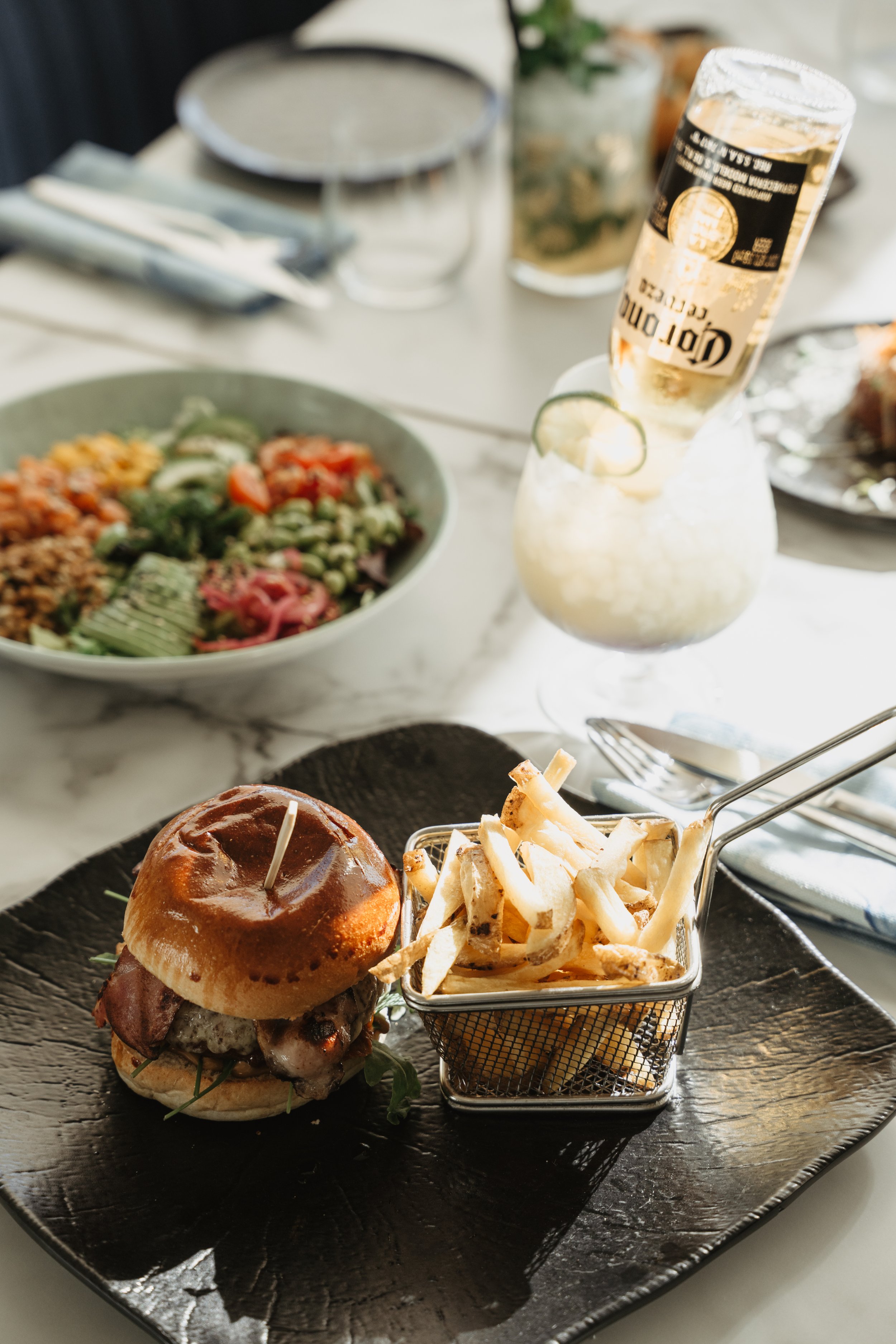 A burger with bacon and cheese on a black plate, served with a side of French fries in a small metal basket, on a table set for dining with a salad, drinks, and utensils.
