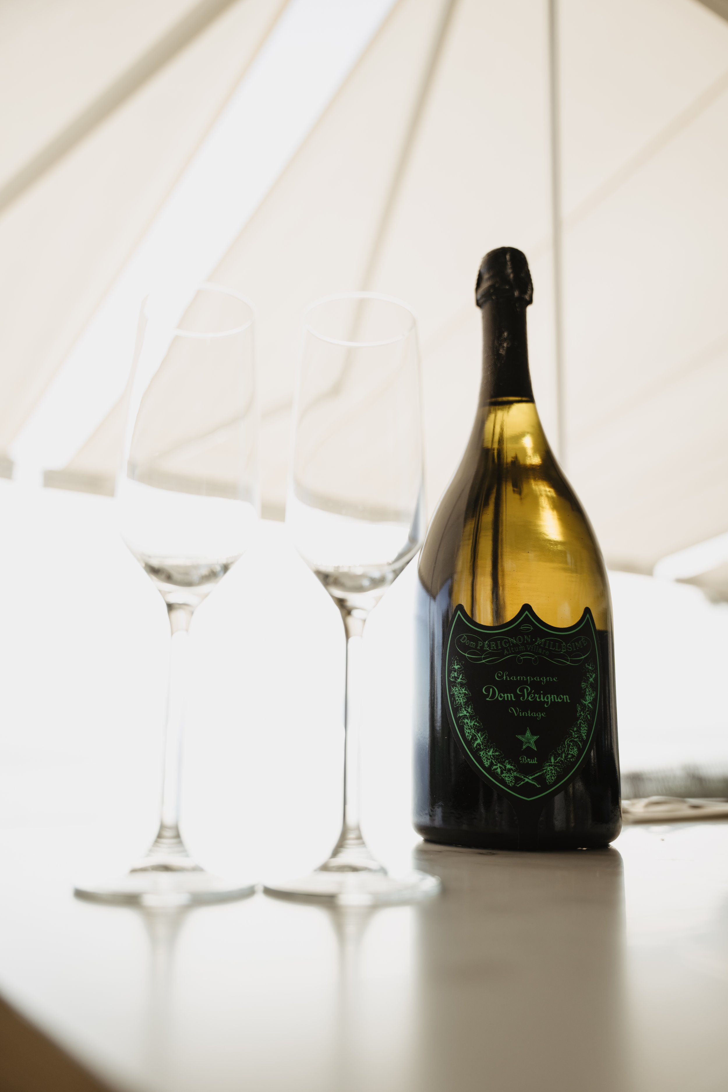 A bottle of Dom Pérignon champagne and two empty champagne glasses on a white surface with a bright background.