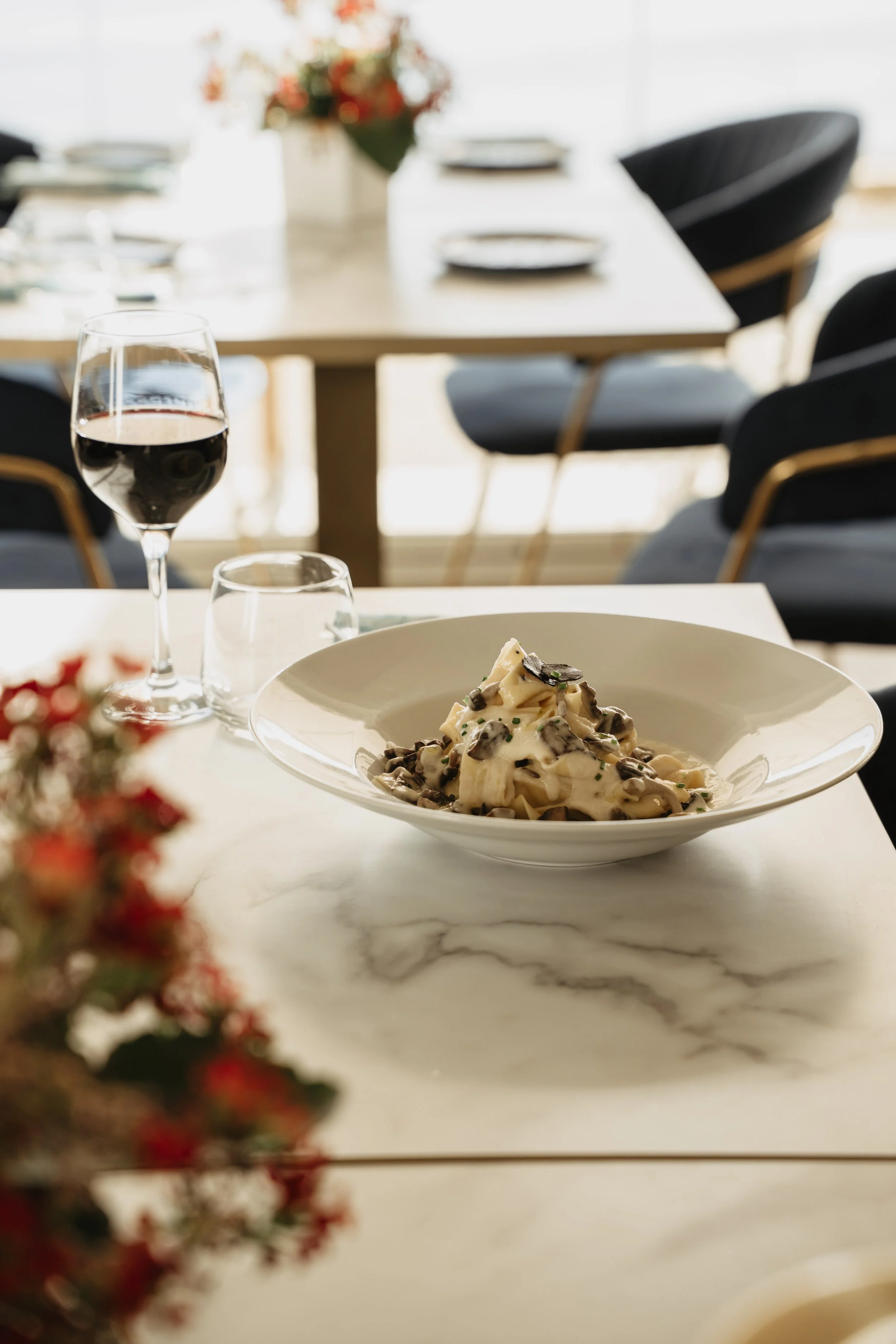 A dining table set with a glass of red wine, a water glass, and a bowl of creamy pasta with mushrooms, with a blurred background of a second table and chairs.
