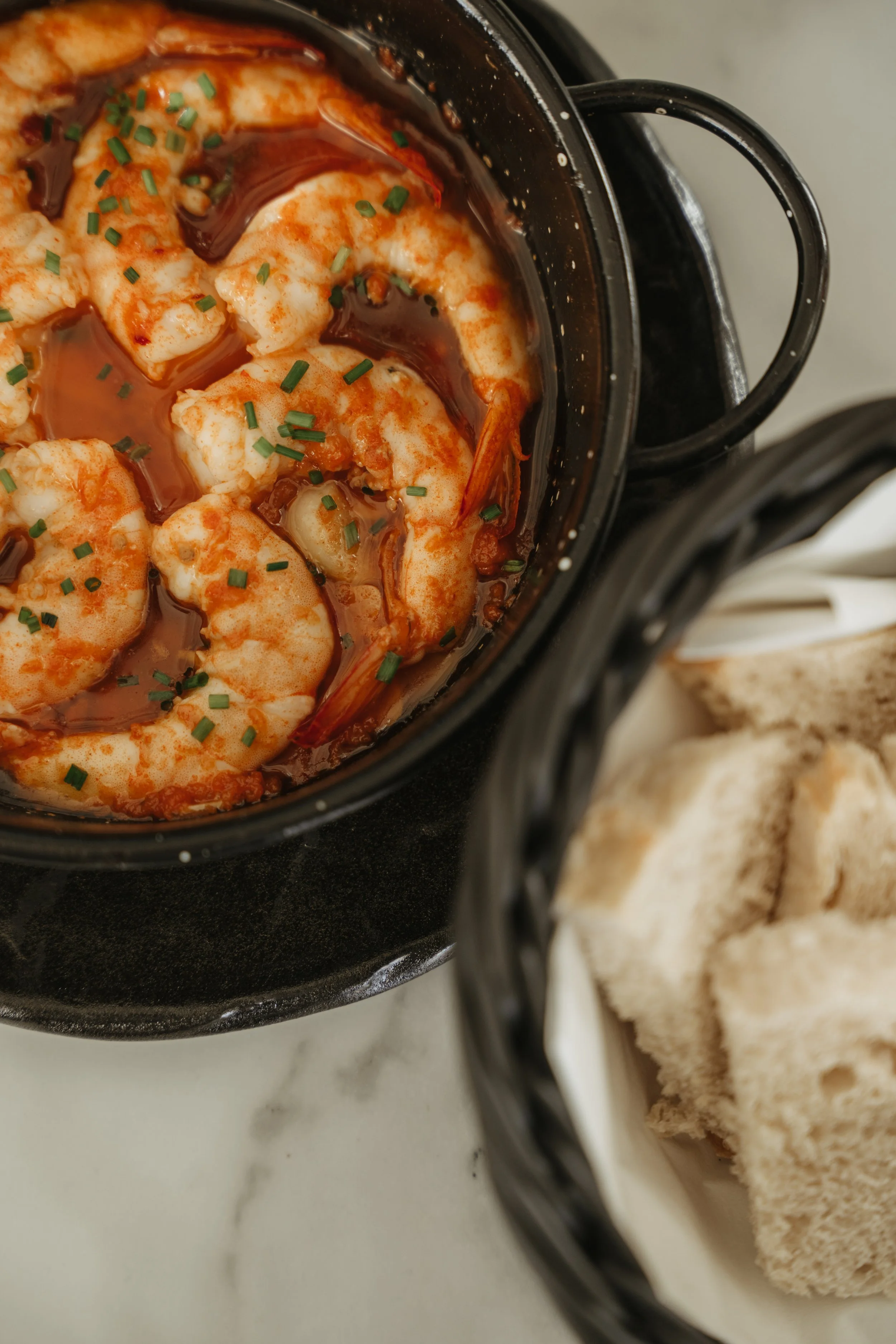 A black bowl of cooked shrimp in a spicy sauce topped with chopped chives, with a side of sliced bread in a basket.