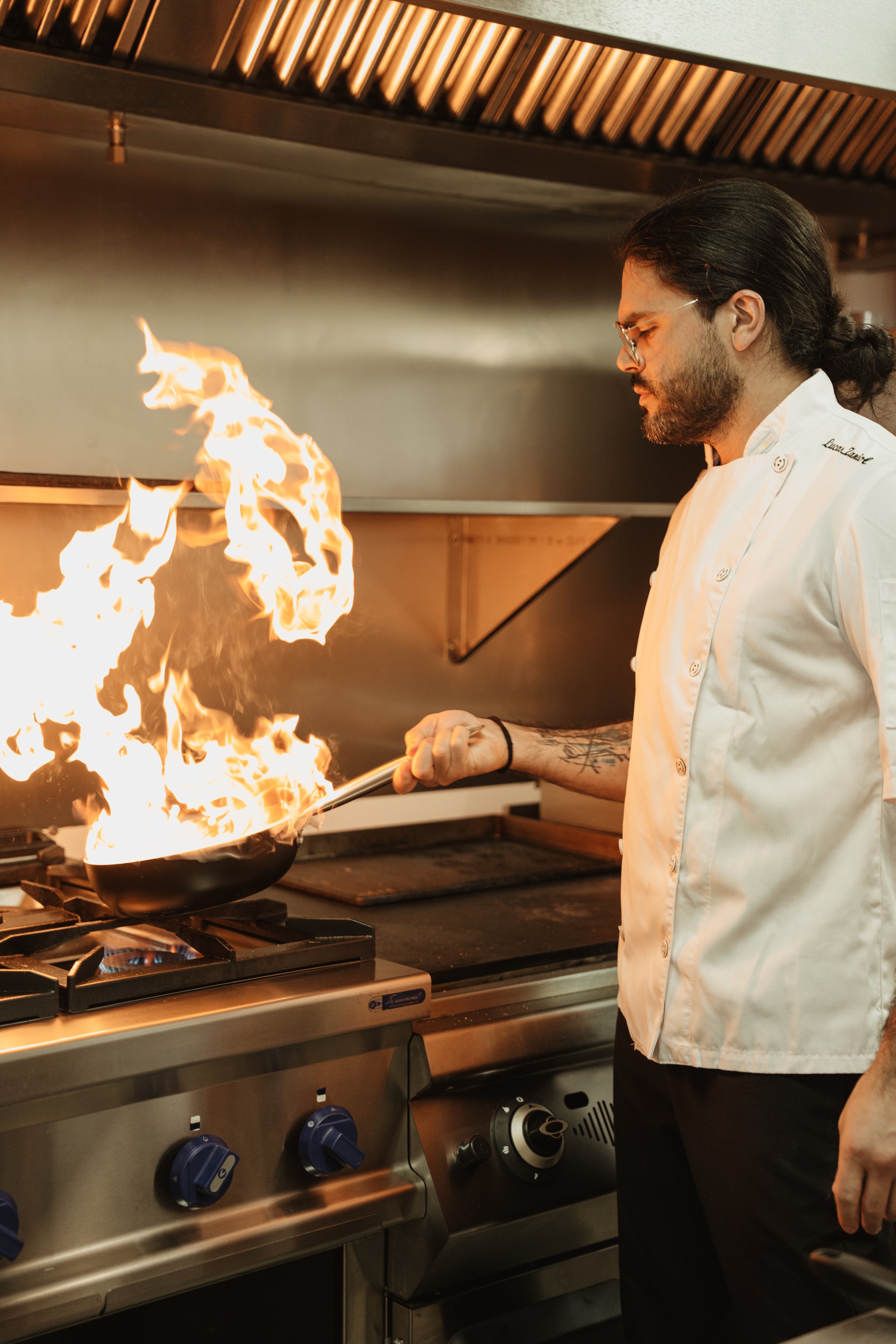 Male chef cooking with a flaming pan in a commercial kitchen.