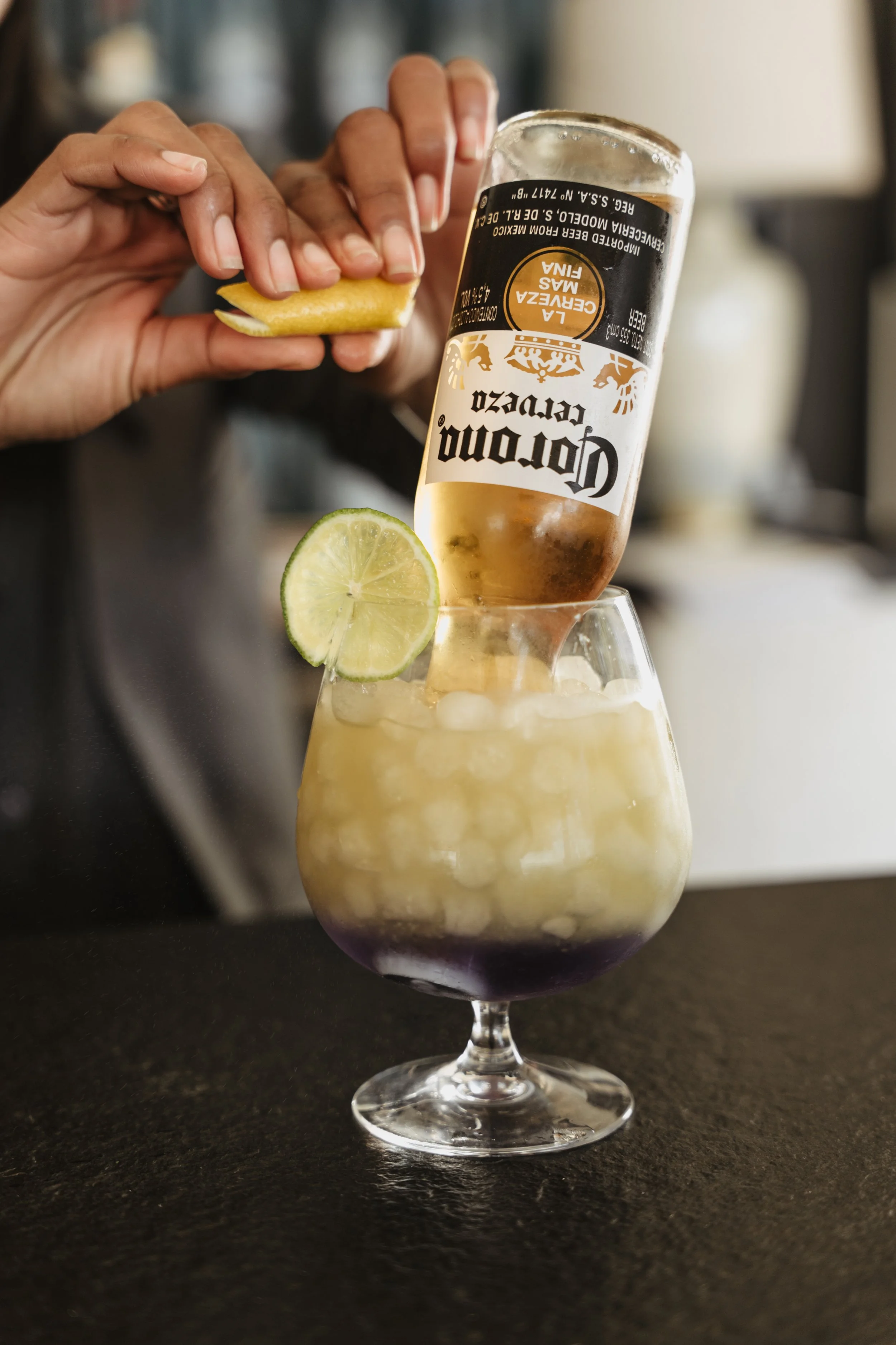 A person is garnishing a cocktail with a lemon wedge while pouring a bottle of Corona beer into the drink, which is in a large glass filled with ice and topped with a lime slice.