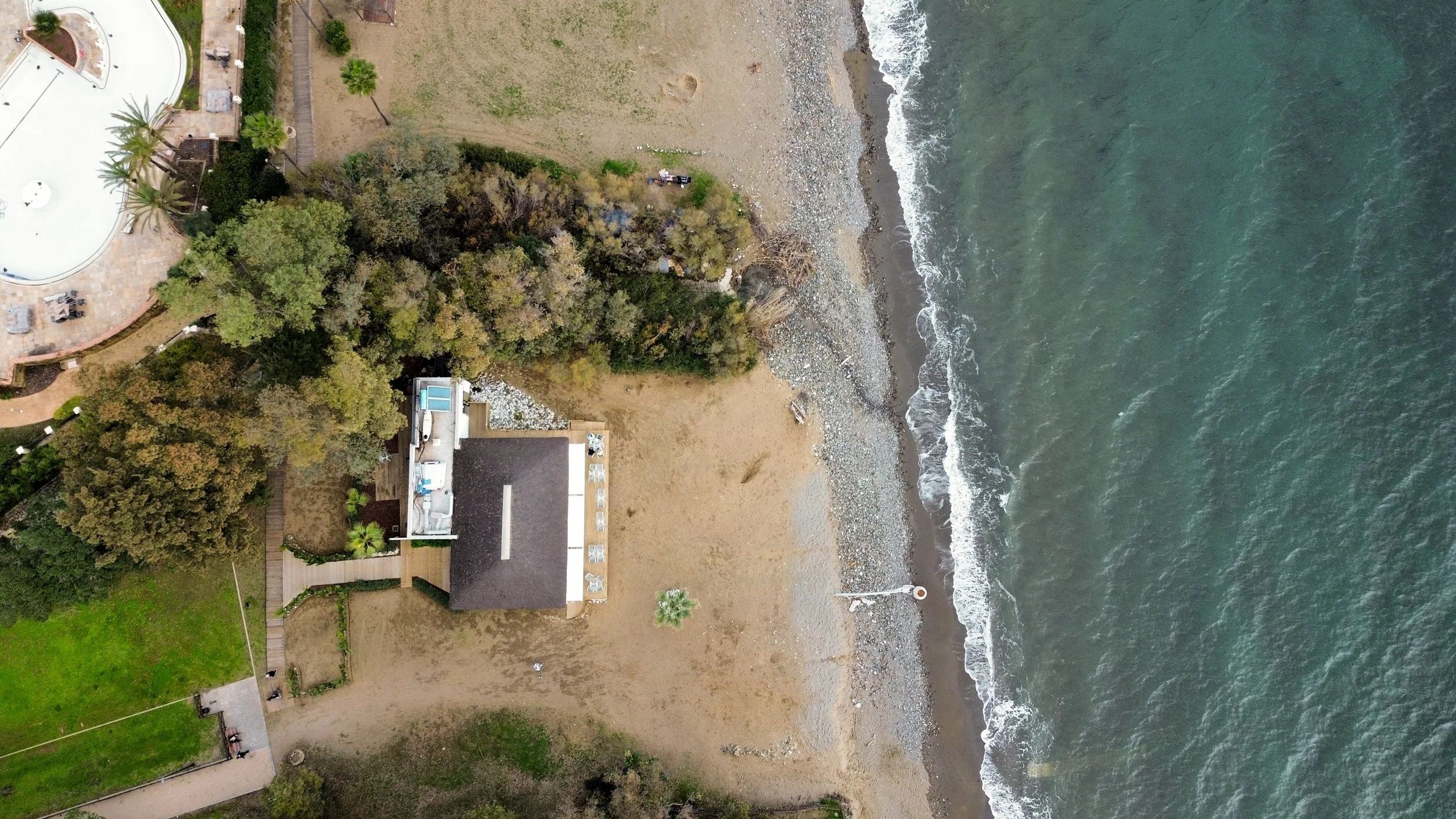 An aerial view of a beach with a house, trees, and a rocky shoreline adjacent to the ocean.