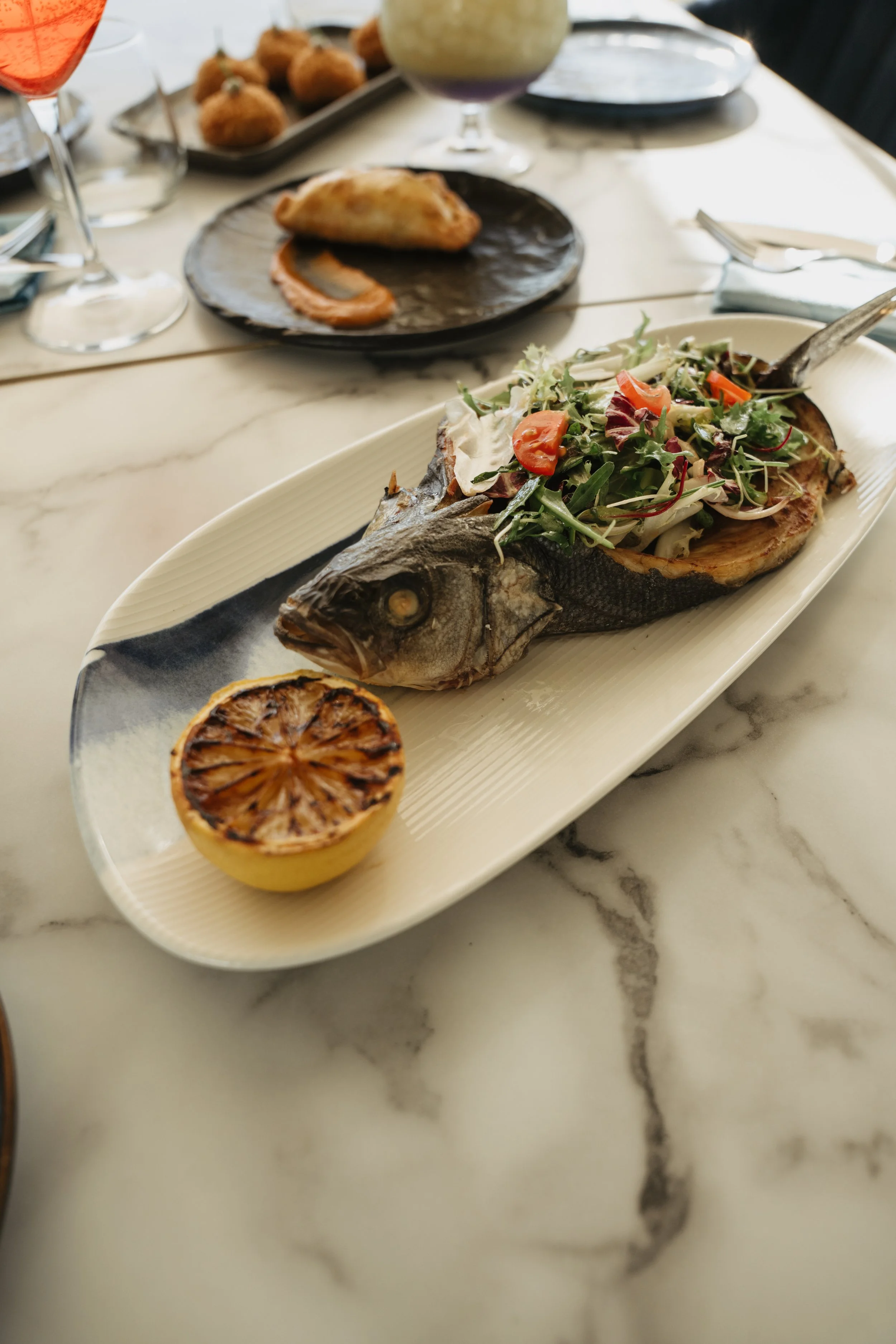 A whole grilled fish served on a white platter with a mixed green salad and a grilled lemon half, on a marble table with other dishes and drinks in the background.