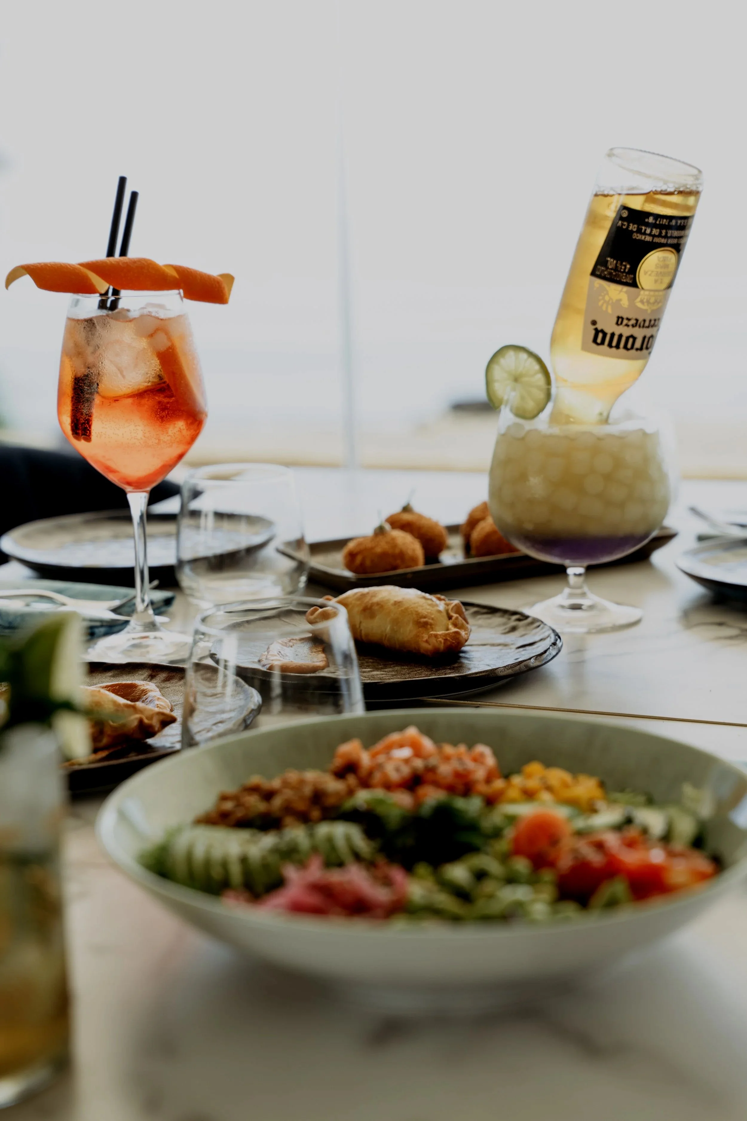 A table set with various drinks and dishes, including a cocktail with orange peel, a beer in a glass, and a salad in a bowl; also features plates with small appetizers and a plate of fried food in the background.